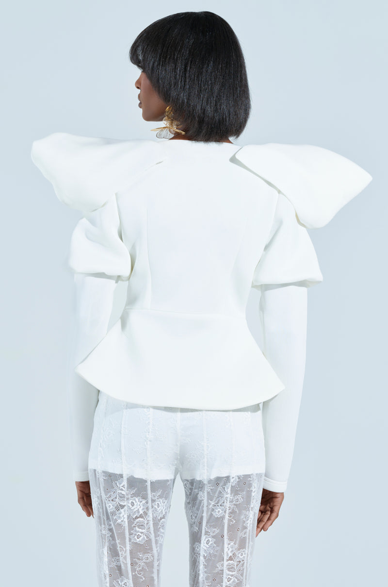 Rear view of someone in a stylish MILA FITTED BLOUSE, featuring structured puffed details and long sleeves. The outfit's elegance shines with sheer lace pants and a plunging v-neckline, set against a light blue background.