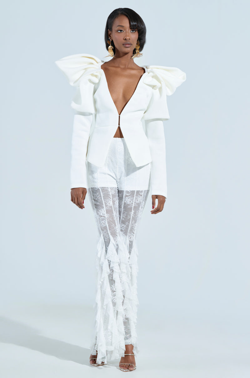 A woman poses in a chic white outfit, with a jacket featuring structured puffed detailing. She pairs elegant lace pants with gold earrings and strappy heels for a sophisticated touch. A MILA FITTED BLOUSE with a plunging v-neckline completes the modern ensemble against a plain background.