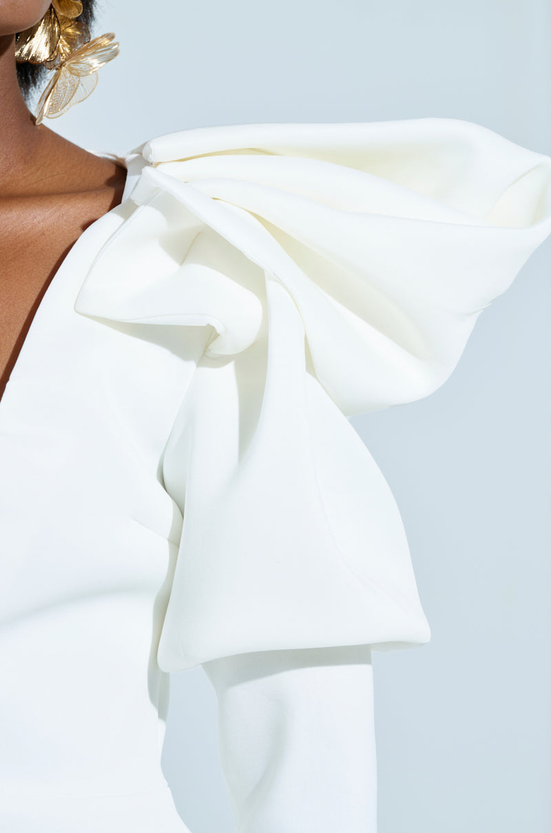 Close-up of a person wearing the MILA FITTED BLOUSE with dramatic, structured puffed shoulders. The elegantly gathered fabric forms a sculptural effect against a soft blue background.