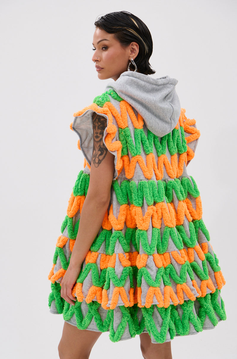 A woman with wet-look dark hair models the LOLO ZIGZAG MINI DRESS in vibrant neon zigzag, featuring ruffled textured fabric and chenille appliqués, plus a gray hood, standing against a plain white background and looking over her shoulder.