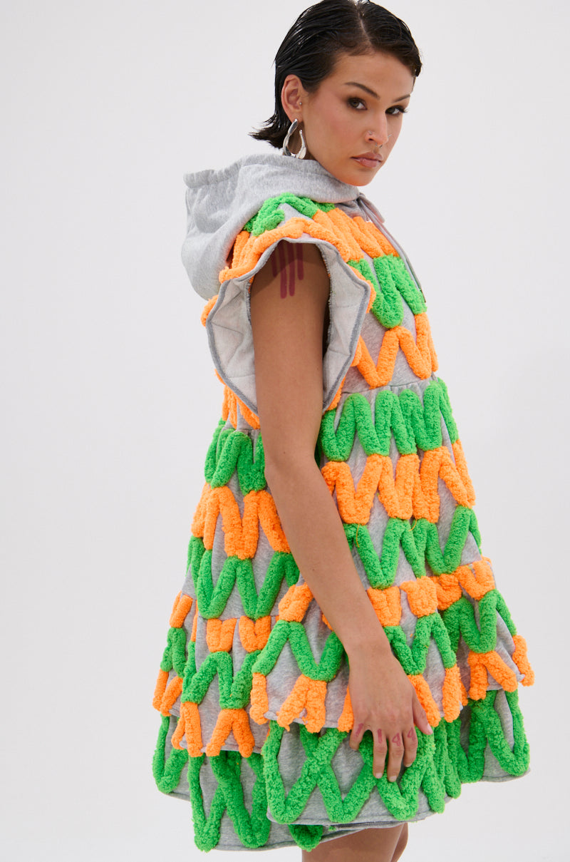 A woman models the LOLO ZIGZAG MINI DRESS, showcasing neon green and orange chenille zigzag appliqués on a gray background. She sports short wet-look hair and large hoop earrings against a plain white backdrop.