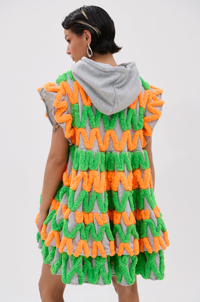 A person with short dark hair wears the LOLO ZIGZAG MINI DRESS, a sleeveless, ruffled mini dress in vibrant neon colors with a gray hood, standing against a plain light background.