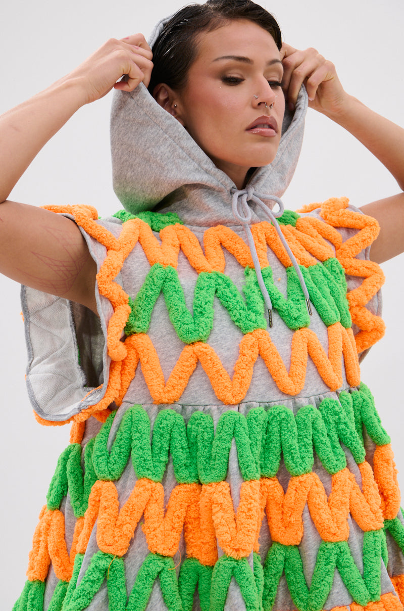 A person with short dark hair and a nose ring pulls up the hood of the LOLO ZIGZAG MINI DRESS, a sleeveless gray mini dress with neon colors and chenille zigzag appliqués. The background is plain and light-colored.
