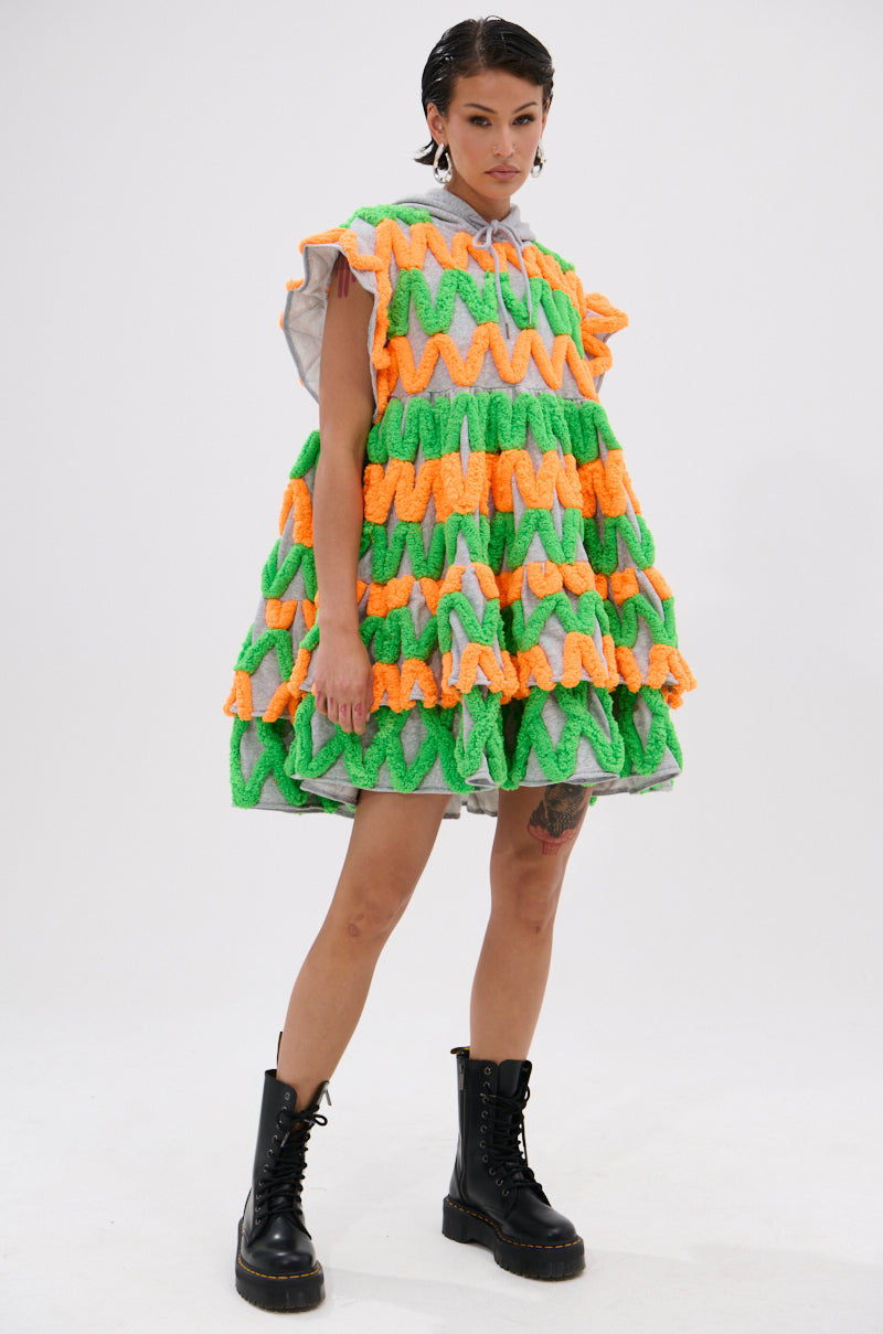 A person stands confidently in the LOLO ZIGZAG MINI DRESS, a loose, sleeveless mini with wavy green, orange, and yellow textured stripes, paired with black lace-up boots against a plain white background.