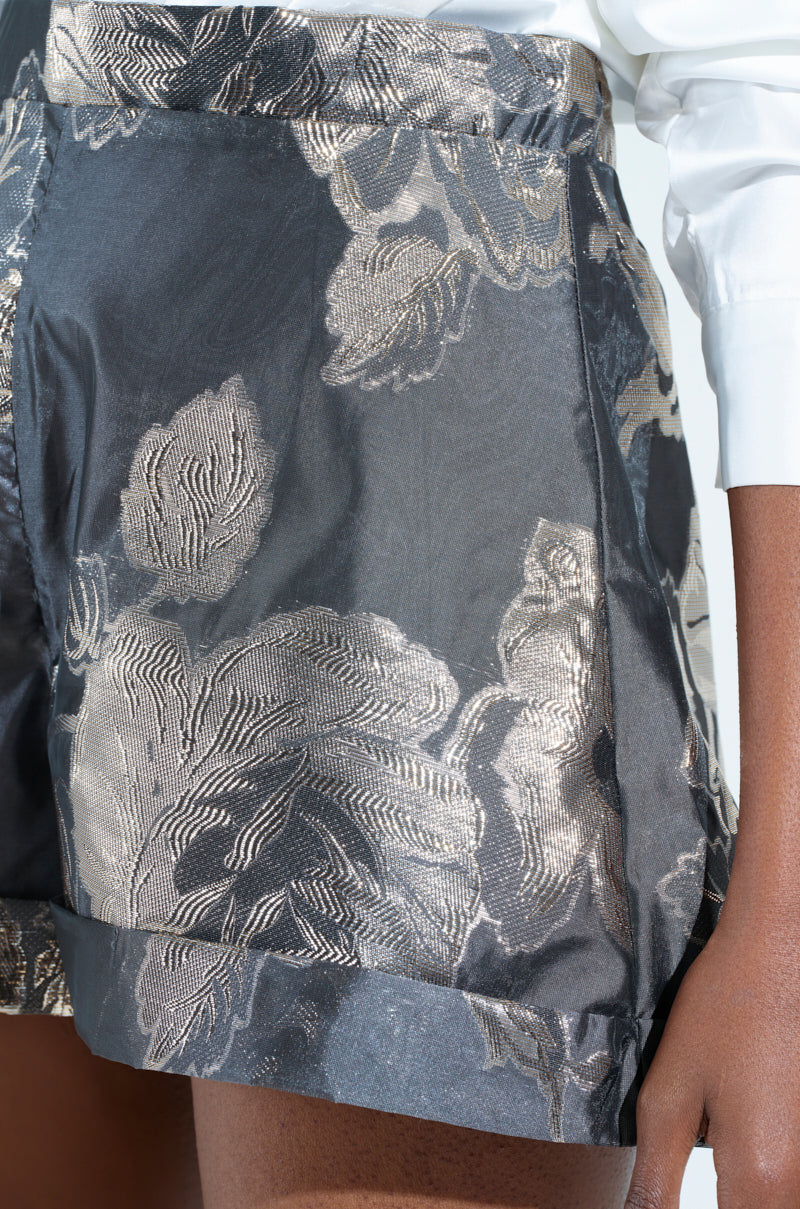 Close-up of a person wearing the DRAMA FLORAL SATIN SHORT, featuring a metallic floral brocade design. The shorts exhibit intricate patterns with a silvery sheen, perfectly paired with a crisp white long-sleeve shirt.
