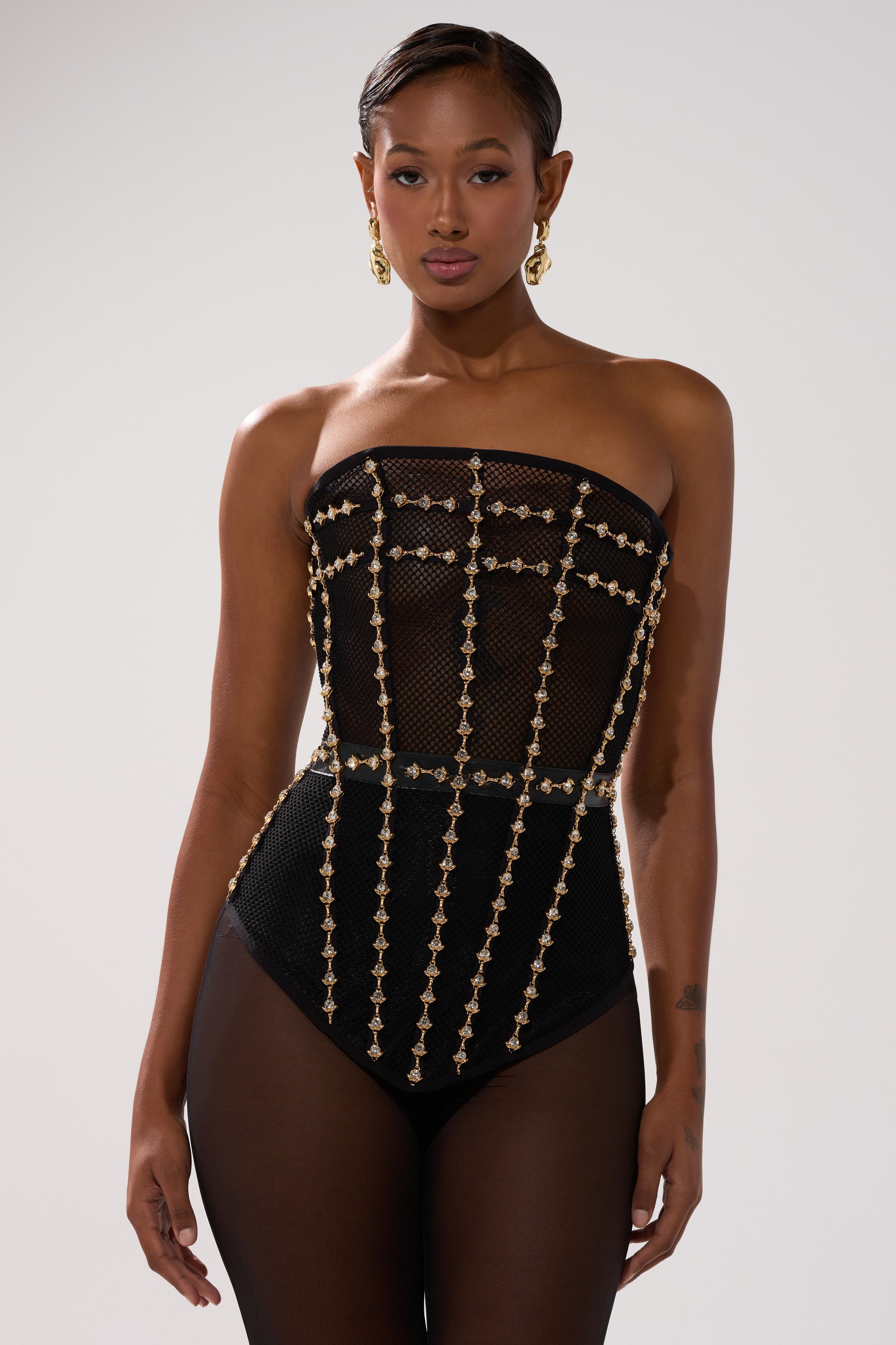 A woman in the SMOKESHOW CORSET WITH RHINESTONES—strapless, sheer mesh with gold chain details—poses confidently against a plain light backdrop, wearing gold hoop earrings and sporting short hair with a neutral expression.