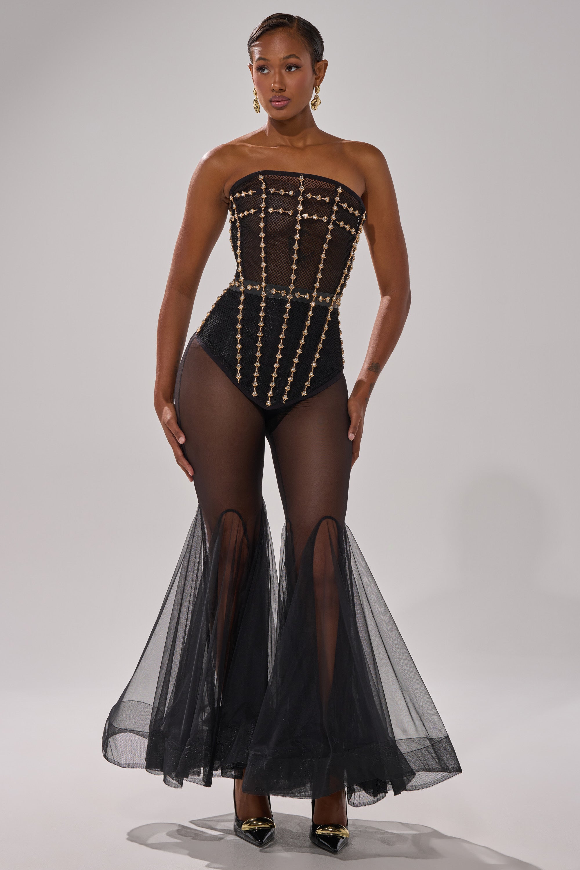 A woman stands confidently in the SMOKESHOW CORSET WITH RHINESTONES, a strapless black sheer mesh bodysuit with rhinestone details, flared tulle pant legs, gold earrings, and black pointed-toe heels against a plain background.