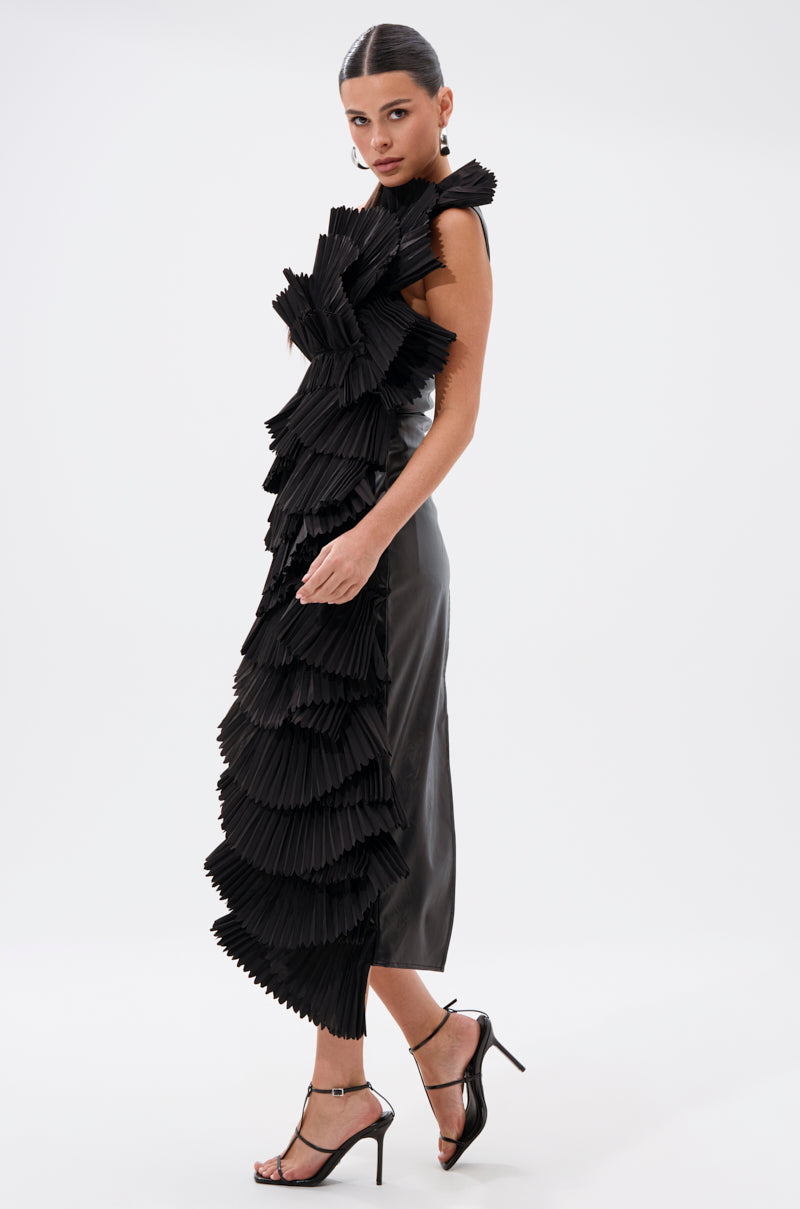 A woman poses in profile against a white background, wearing the LUXURIOUS ASYMMETRICAL TOP IN BLACK styled as a sleeveless midi dress with dramatic layered pleated ruffles down one side, paired with black high-heeled sandals and slicked-back hair.