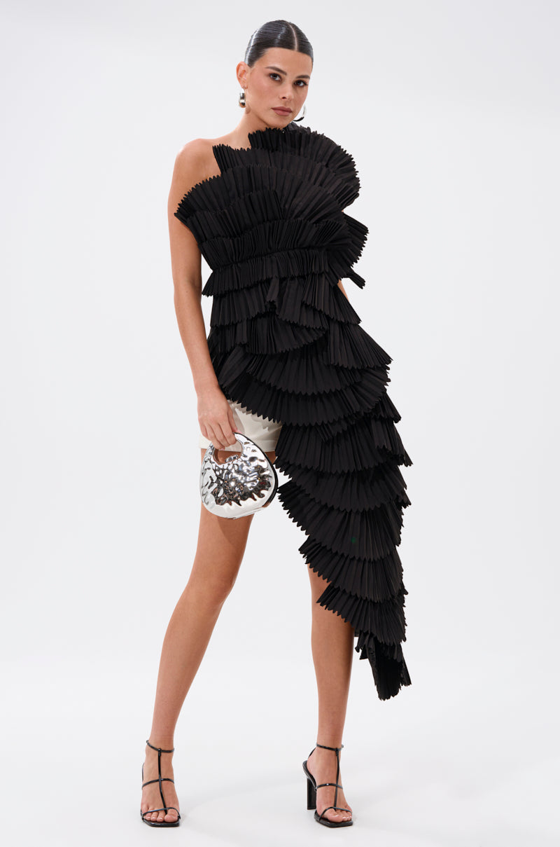 A woman poses against a plain background wearing the LUXURIOUS ASYMMETRICAL TOP IN BLACK, featuring dramatic pleated ruffles and one exposed shoulder. She pairs it with black strappy heels, a silver handbag, and a sleek, tied-back hairstyle.
