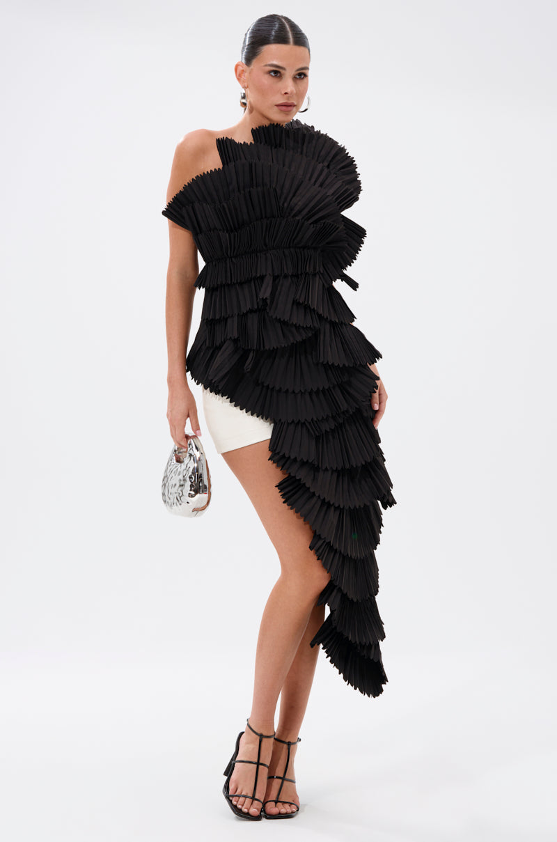 A woman poses against a plain white background wearing the LUXURIOUS ASYMMETRICAL TOP IN BLACK with pleated ruffles, styled over white shorts, and accessorized with a patterned purse and strappy black heels.