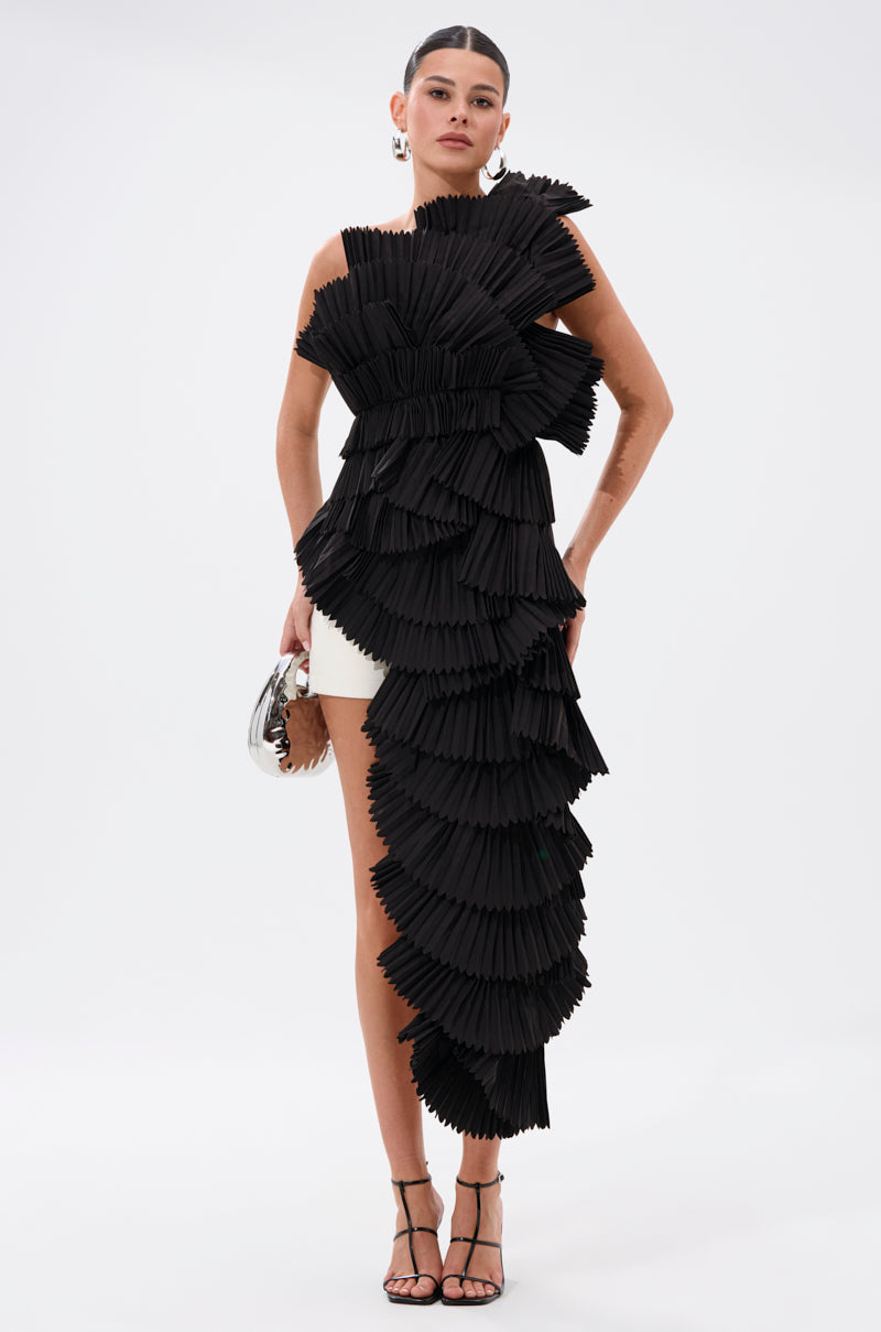 A woman stands against a plain background wearing the LUXURIOUS ASYMMETRICAL TOP IN BLACK, featuring unique pleated ruffle and dramatic asymmetrical details, paired with a short white skirt, silver clutch, and high-heeled sandals.