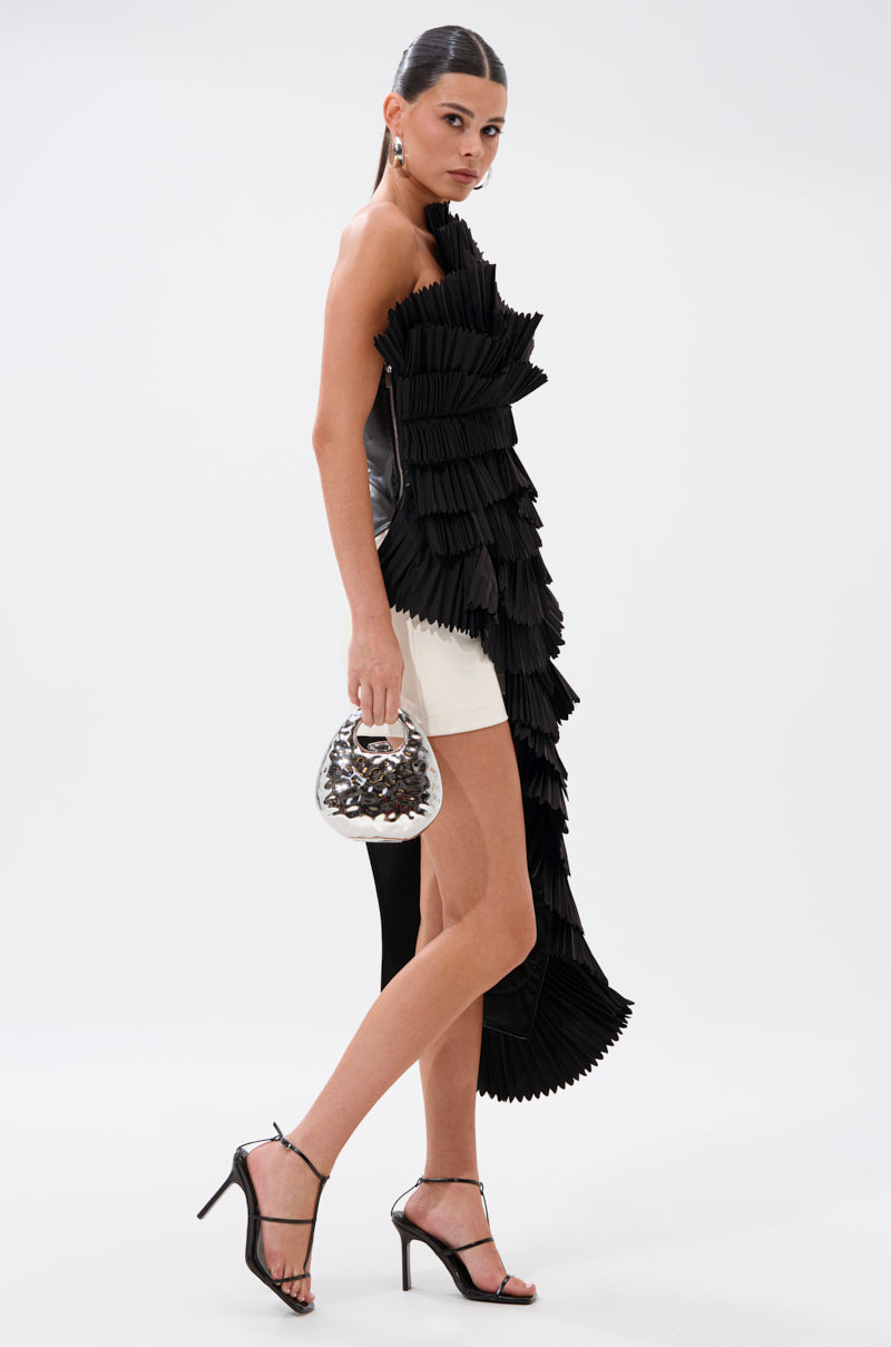 A woman in profile wears the LUXURIOUS ASYMMETRICAL TOP IN BLACK with pleated ruffles, paired with a short white skirt, black high-heeled sandals, a shiny silver handbag, and her hair pulled back.