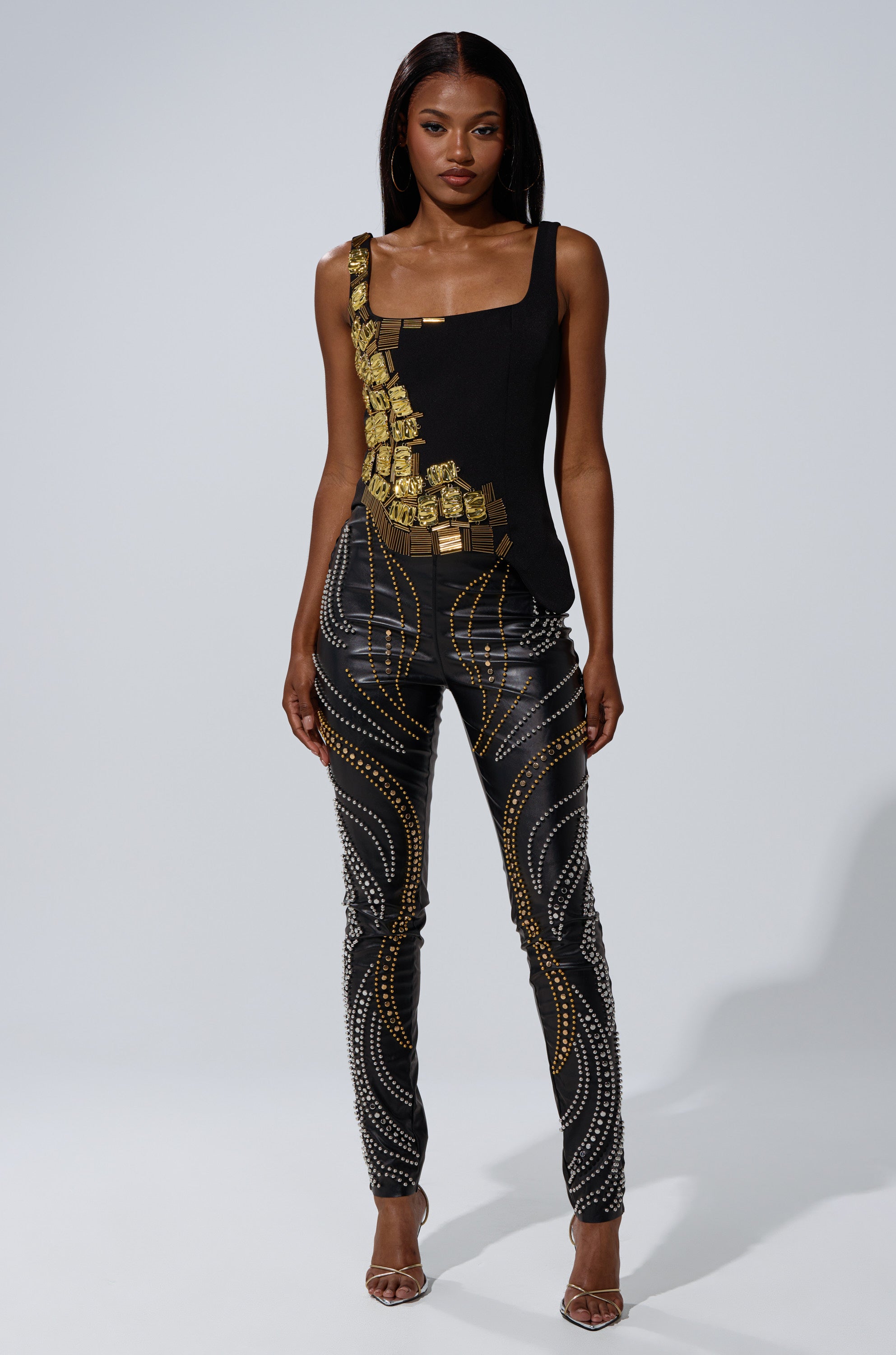 A woman stands against a plain background wearing the HAND BEADED LUX BLOUSE with gold hardware accents and fitted black pants adorned with gold and silver dotted patterns, finishing the look with strappy heels.