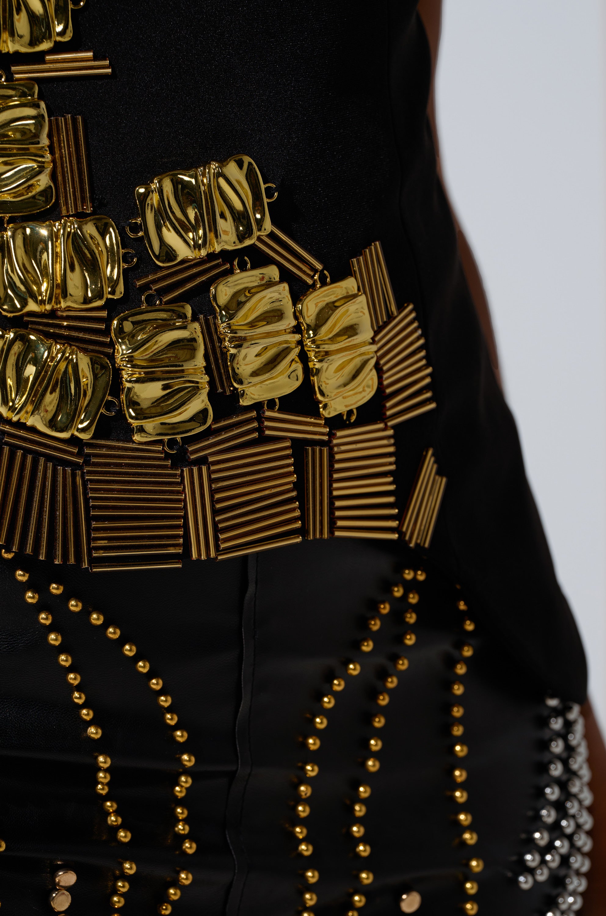 Close-up of the HAND BEADED LUX BLOUSE, featuring an asymmetrical black design with shiny gold geometric embellishments, metallic studs, and ornate gold hardware accents that highlight the blouse’s rich textures and detail.
