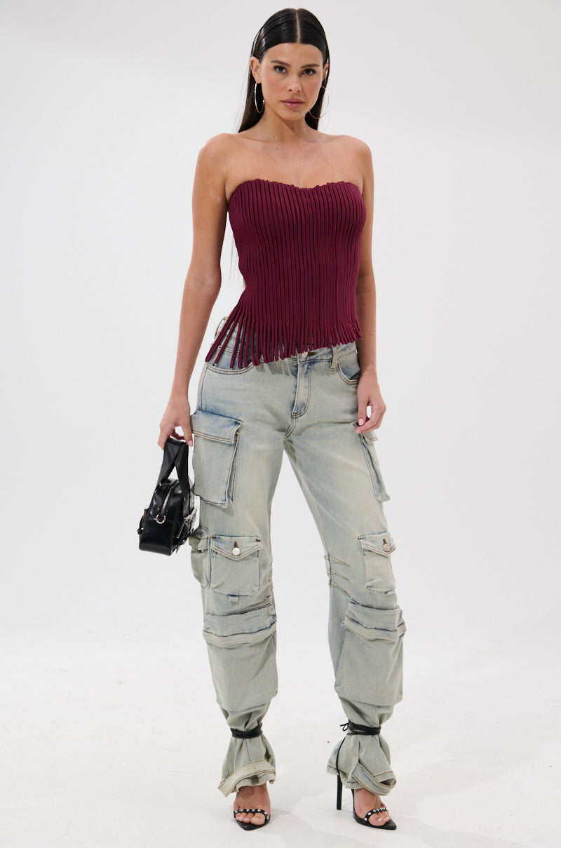 A woman models the ACCORDION CORSET TOP IN BURGUNDY with a pleated overlay and fringed hem, paired with light wash cargo jeans, black strappy heels, and a small black handbag. Her straight dark hair is styled in a center part.