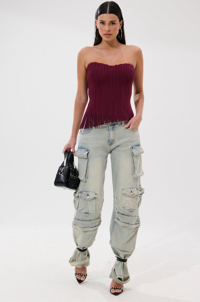 A woman stands against a white background wearing the Accordion Corset Top in Burgundy, light-wash cargo jeans with tied ankles, black heeled sandals, hoop earrings, and carrying a small black handbag.