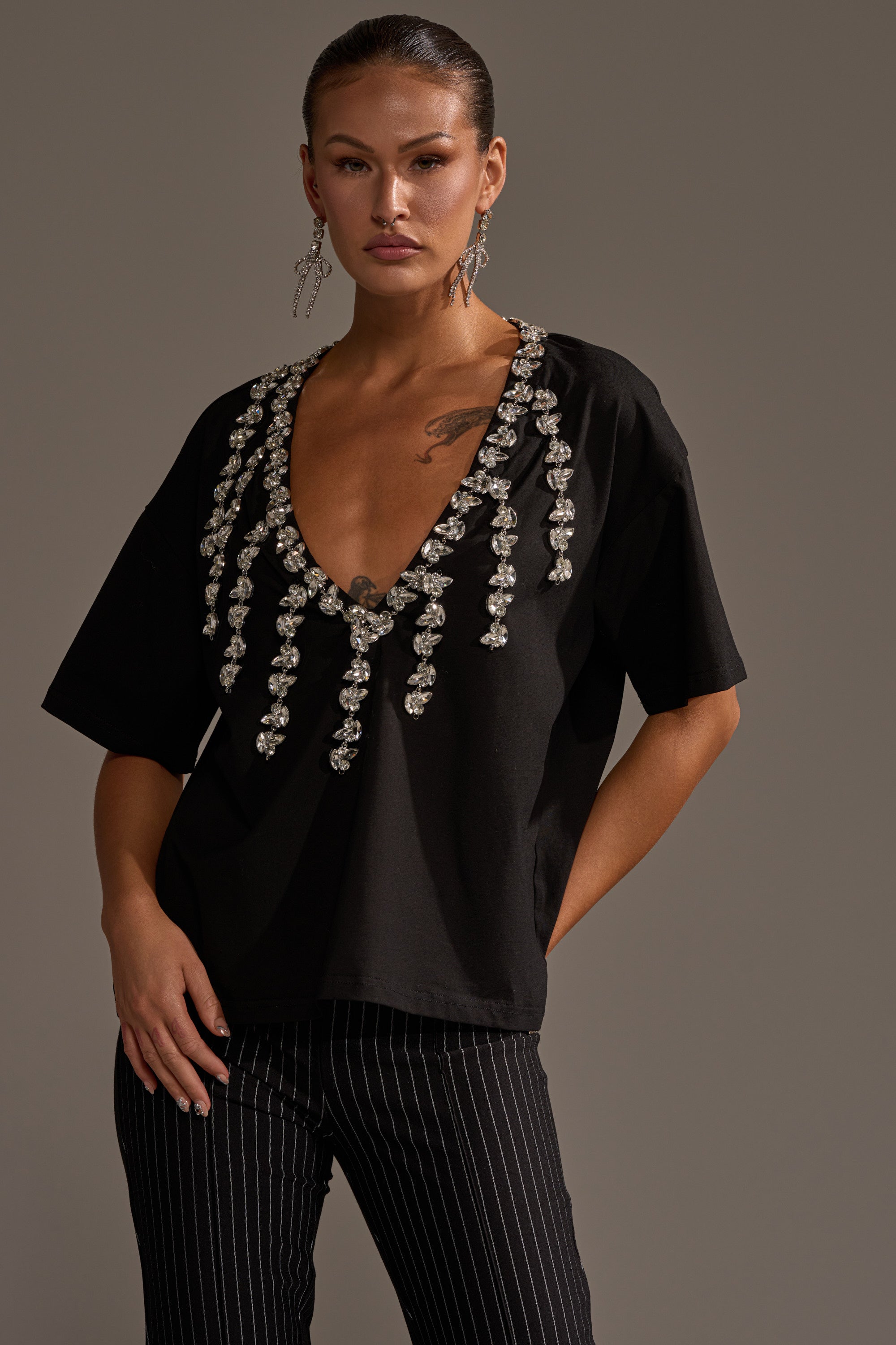 Wearing the RHINESTONE QUEEN OVERSIZED TEE with a plunging V-neck and hanging bead embellishments, paired with black pinstriped pants. Slick-back hair and long, dangling earrings complete the sophisticated look against a plain background.