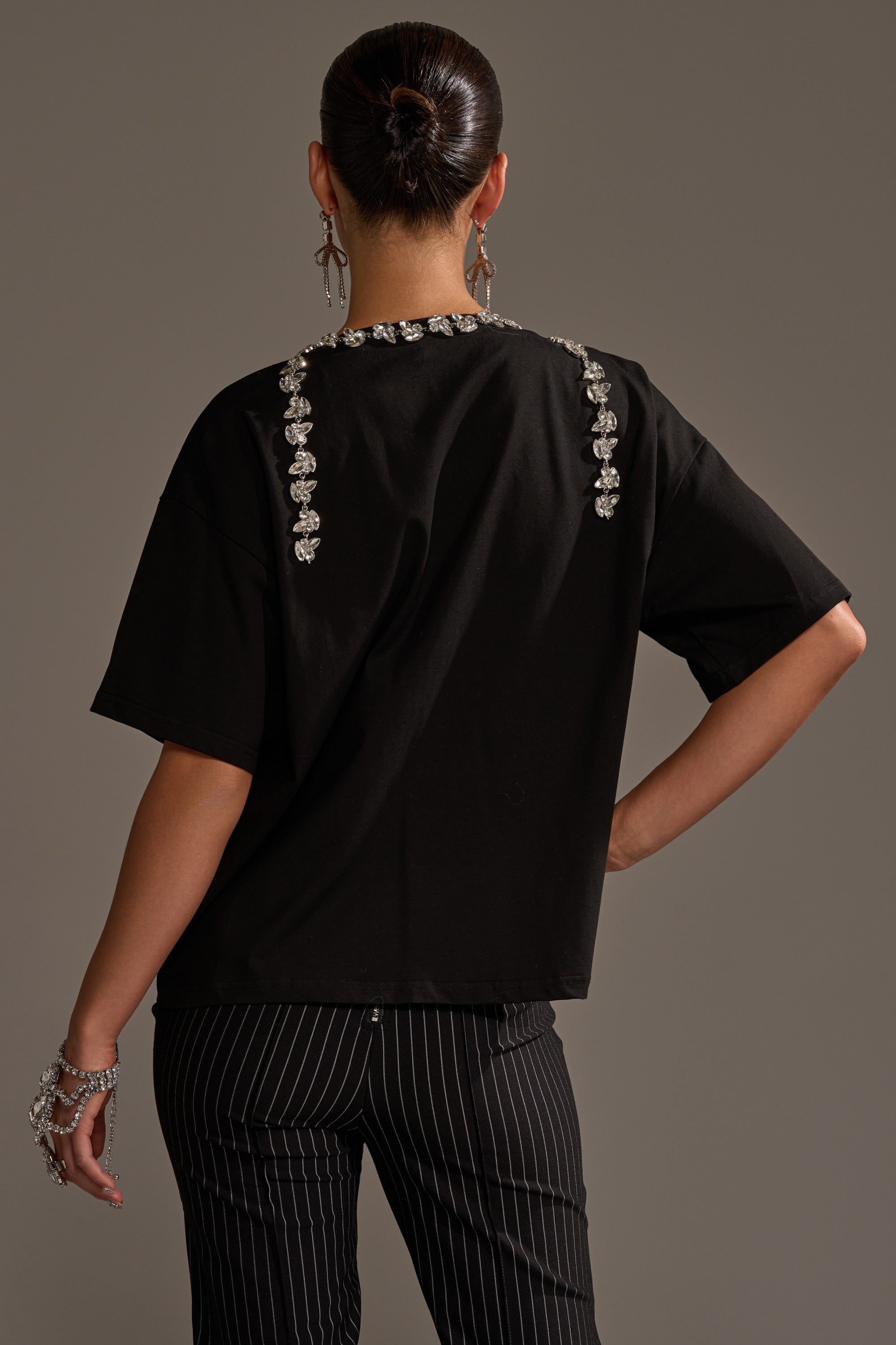 A person faces away, wearing the RHINESTONE QUEEN OVERSIZED TEE with jeweled details on the shoulders and back seam, styled with black pinstripe pants, a neat bun, statement earrings, and a bracelet.