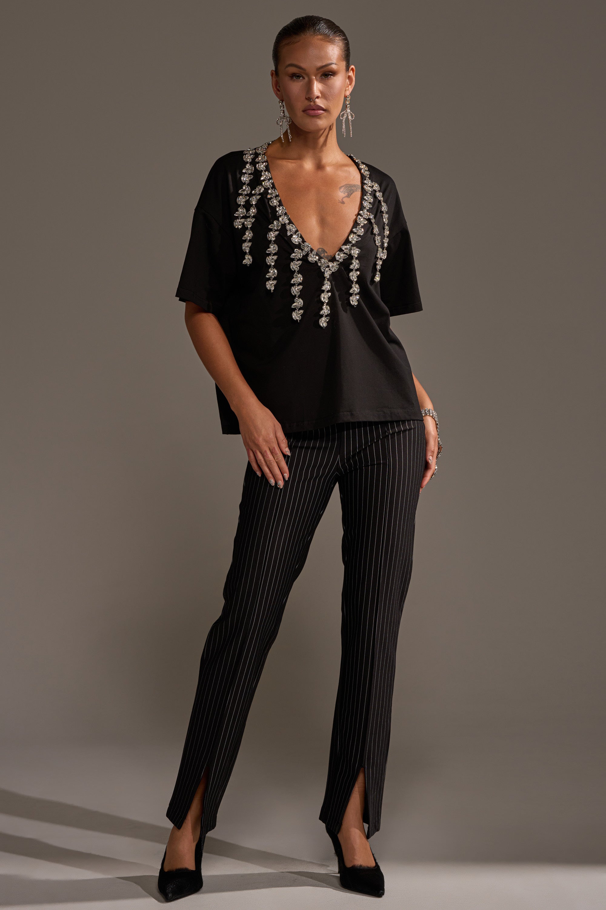 A person stands confidently in the RHINESTONE QUEEN OVERSIZED TEE with a plunging V-neckline and hanging embellishments, paired with black pinstripe pants with front slits, black heels, and statement earrings against a plain background.