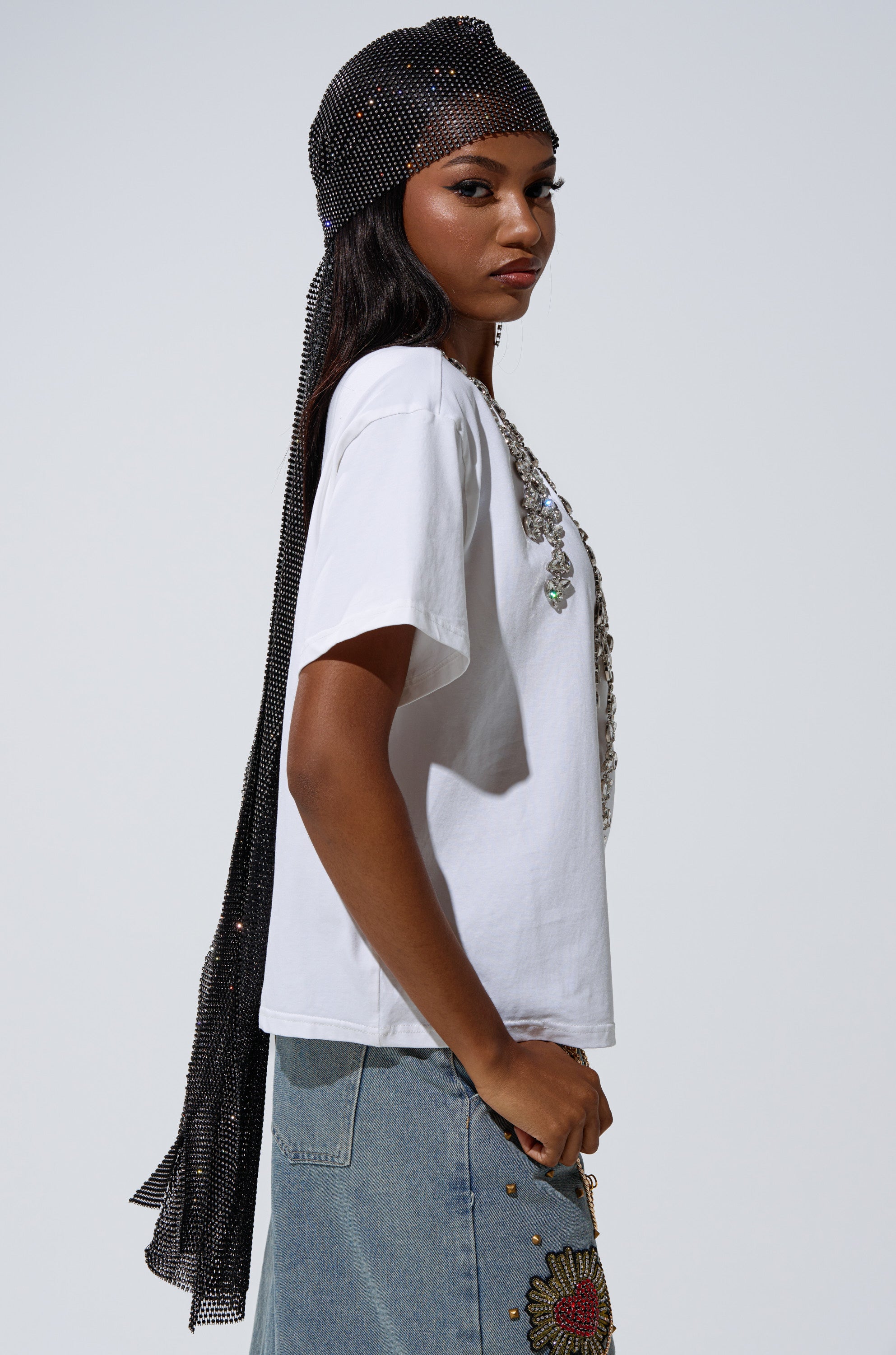 A woman poses in profile wearing the TOO DEEP RHINESTONE TEE, a light blue embroidered denim skirt, a sparkly black headscarf draped down her back, and a statement necklace over her long dark hair.