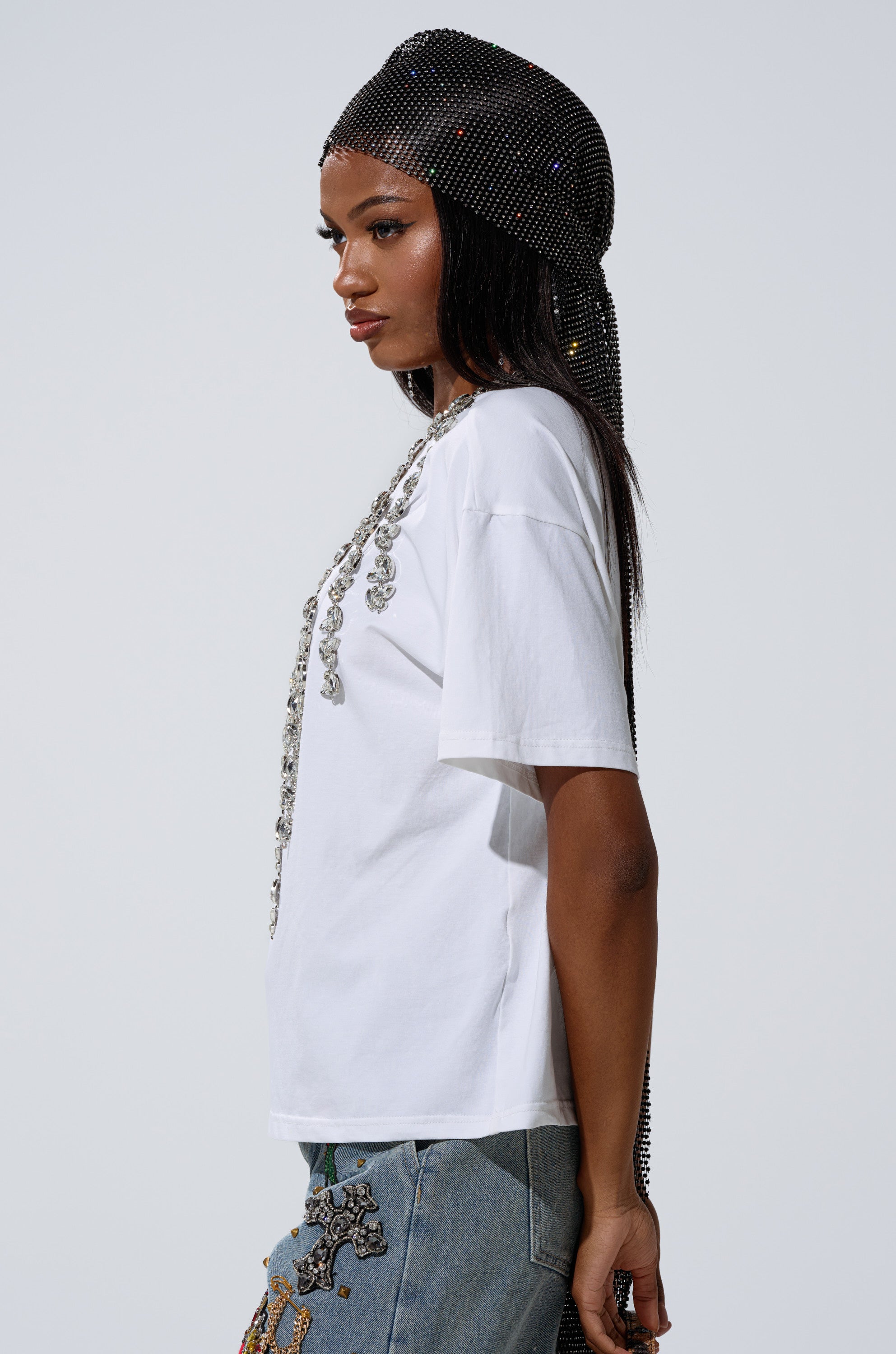 A woman poses in profile wearing the TOO DEEP RHINESTONE TEE with embellished jeans and a black rhinestone-studded headscarf against a plain light background.