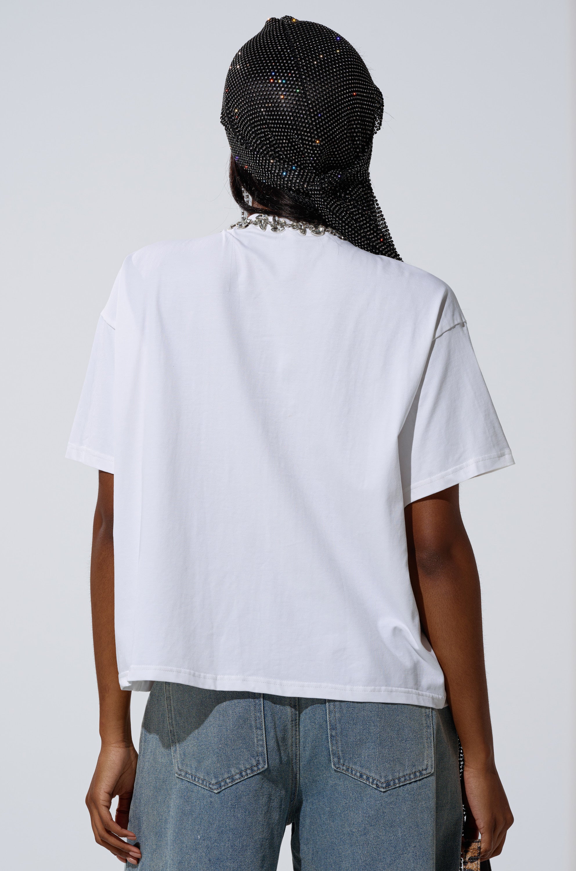 Wearing the TOO DEEP RHINESTONE TEE, light blue jeans, and a sparkling black headscarf, a person stands with their back to the camera against a plain background.