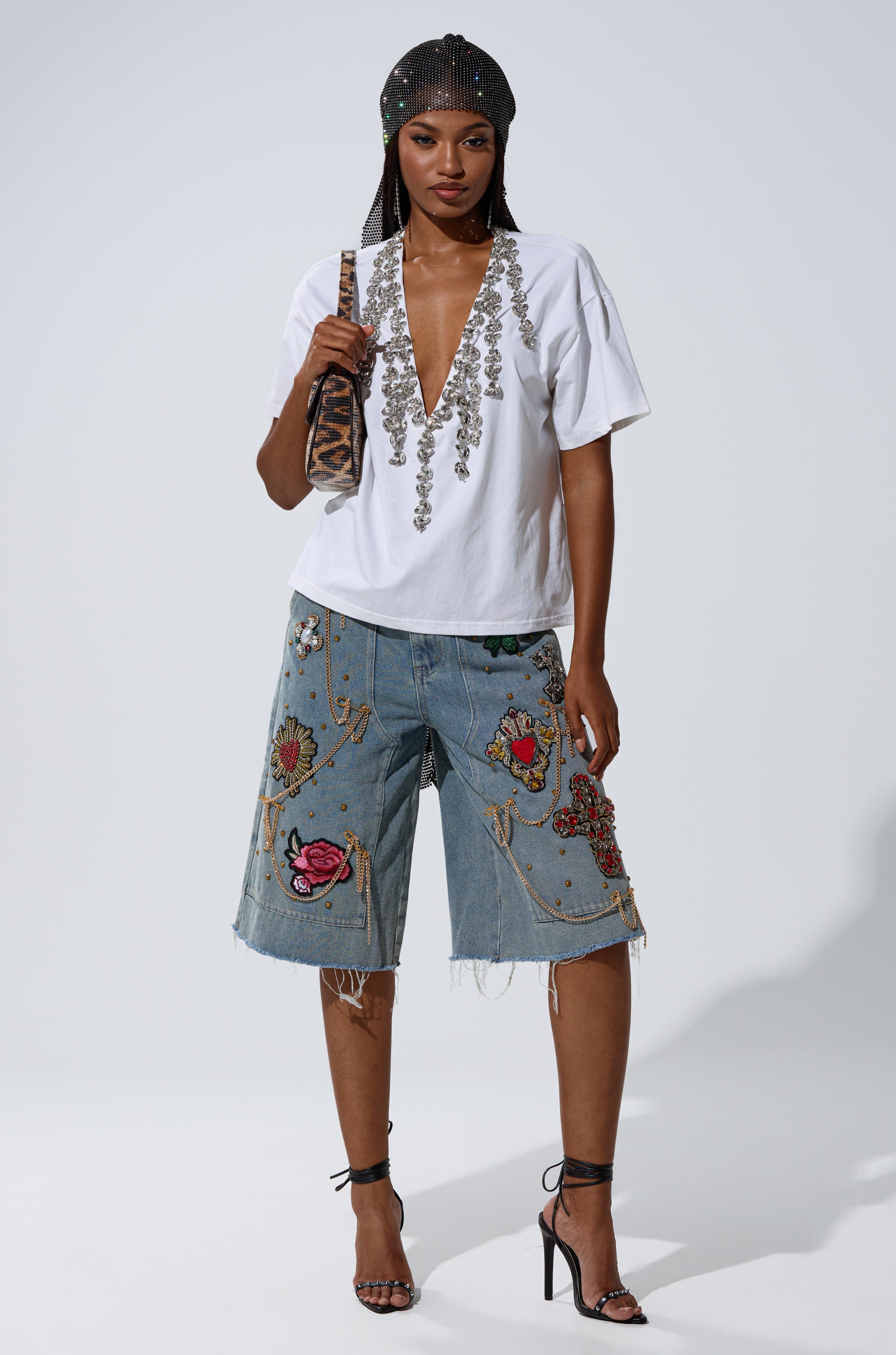 A woman stands confidently in the TOO DEEP RHINESTONE TEE with a plunging v-neckline, paired with embroidered denim knee-length shorts, strappy black heels, a beaded headscarf, and an animal-print shoulder bag.