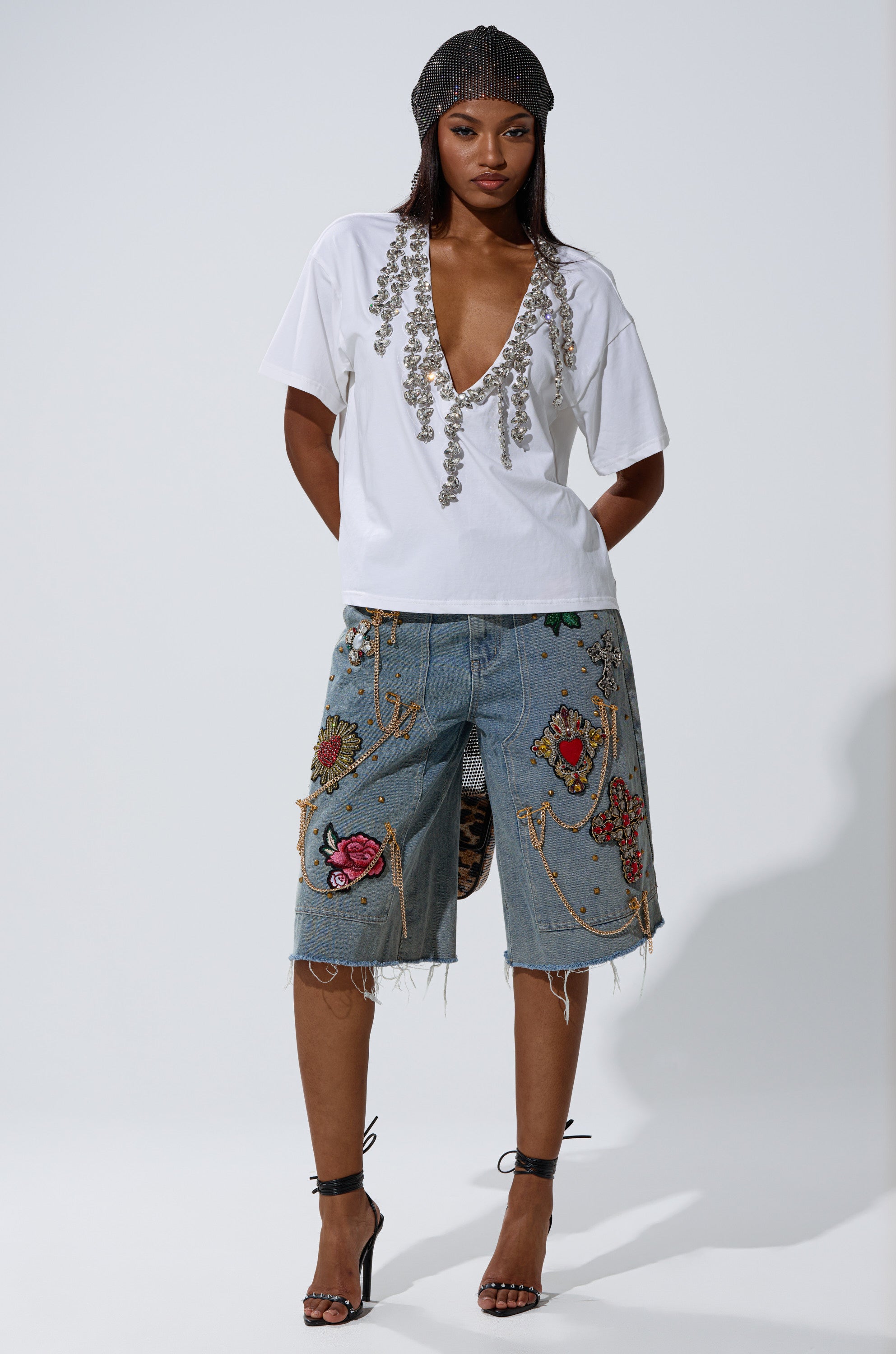 A woman wears the TOO DEEP RHINESTONE TEE with a plunging v-neckline, embroidered distressed knee-length denim shorts, strappy black heels, and a mesh headscarf against a plain light background.