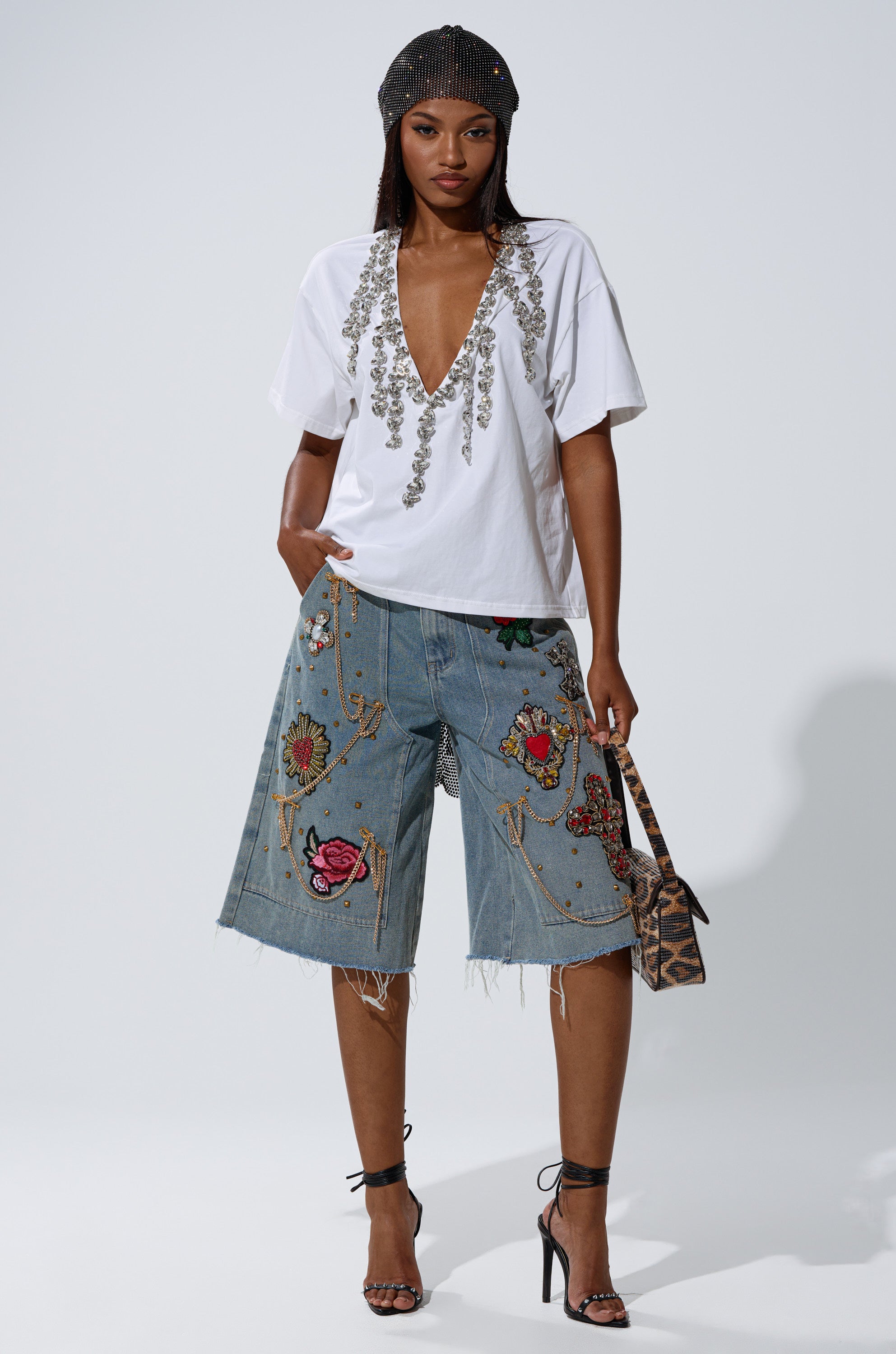 A woman in the TOO DEEP RHINESTONE TEE with a plunging v-neck, beaded black headscarf, and embroidered knee-length denim shorts stands confidently in heels, holding a small animal-print handbag.