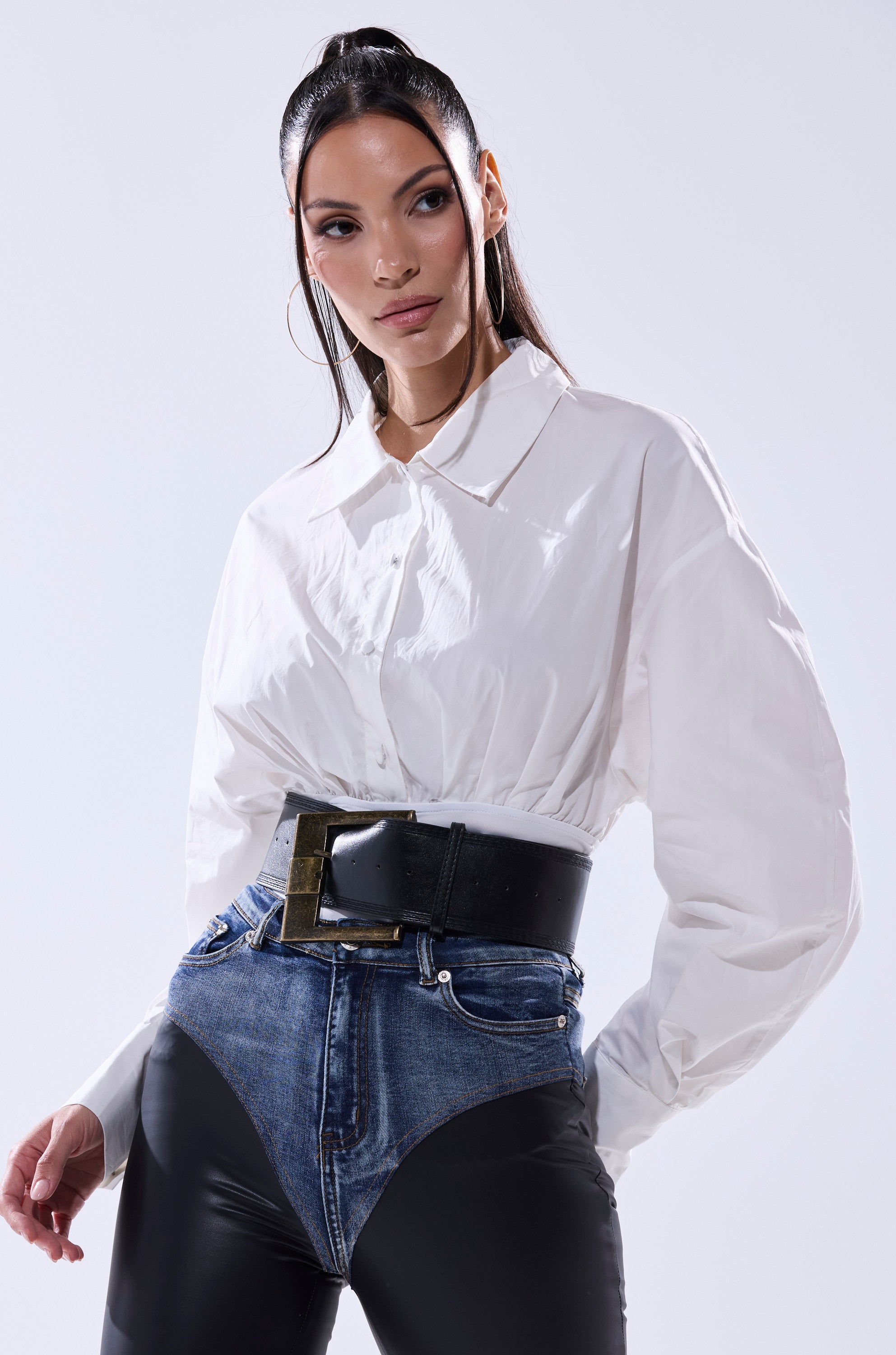 A woman with long dark hair in a high ponytail wears the LET'S GO BABY BODYSUIT, paired with a wide belt and high-waisted pants featuring denim and black leather panels, standing against a plain light background.