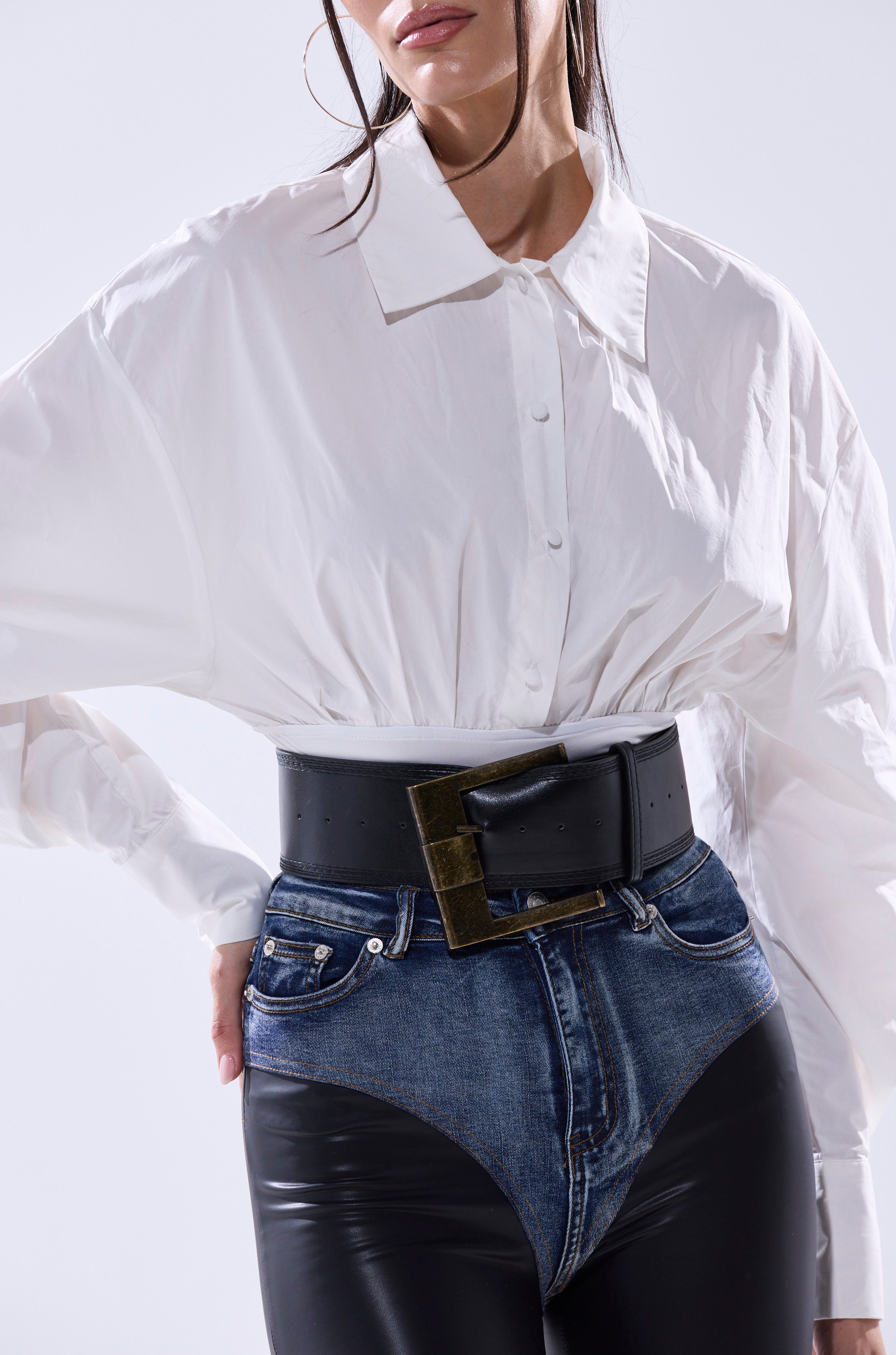 A woman wears the LET'S GO BABY BODYSUIT, paired with high-waisted denim pants featuring black leather-like panels and a wide faux leather belt with a large rectangular buckle. Her face is partially out of frame.