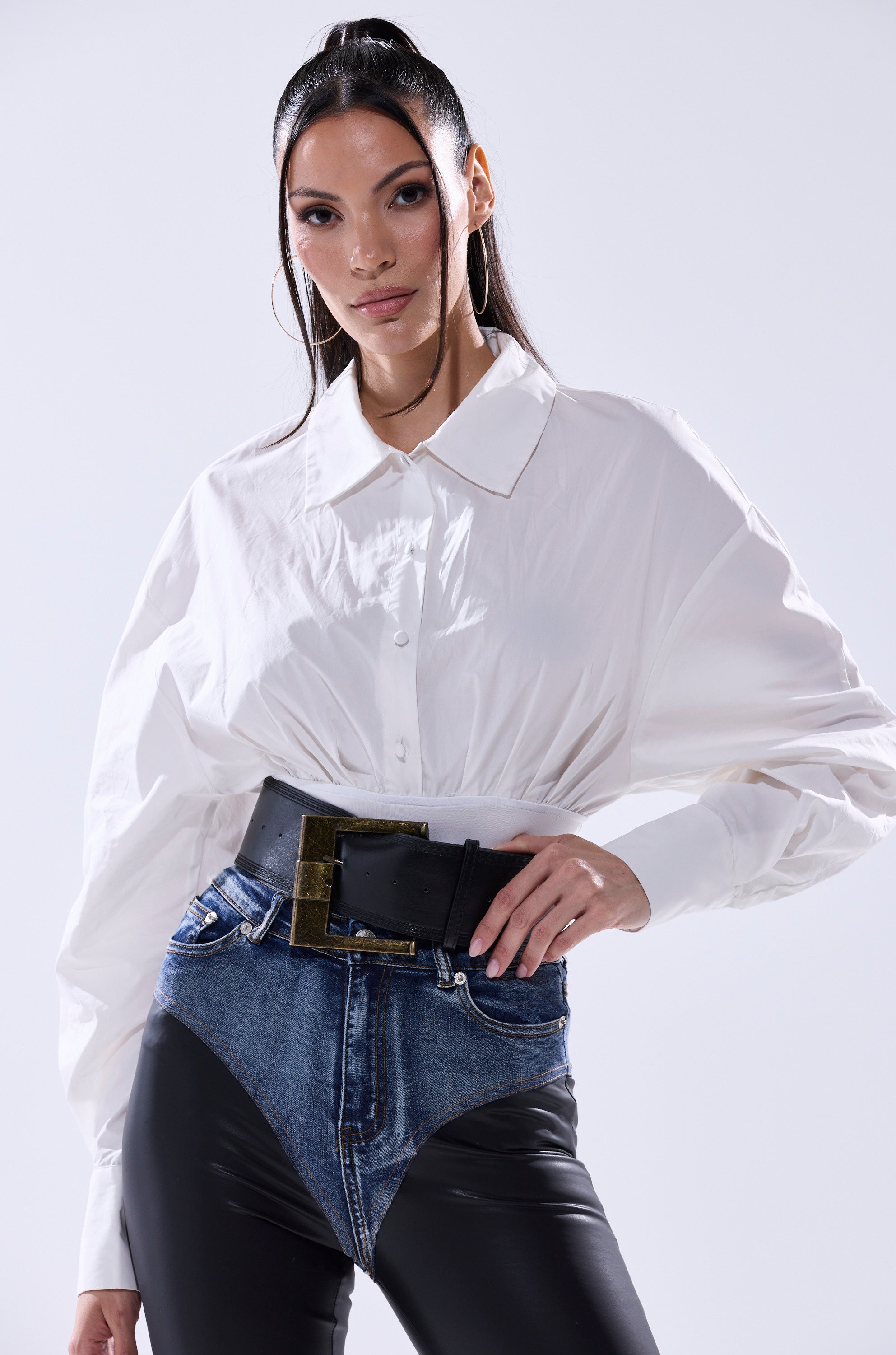 A woman poses confidently in the LET'S GO BABY BODYSUIT—a white collared style with wide sleeves—paired with large hoop earrings, high-waisted jeans featuring black leather panels, and a chic faux leather belt.