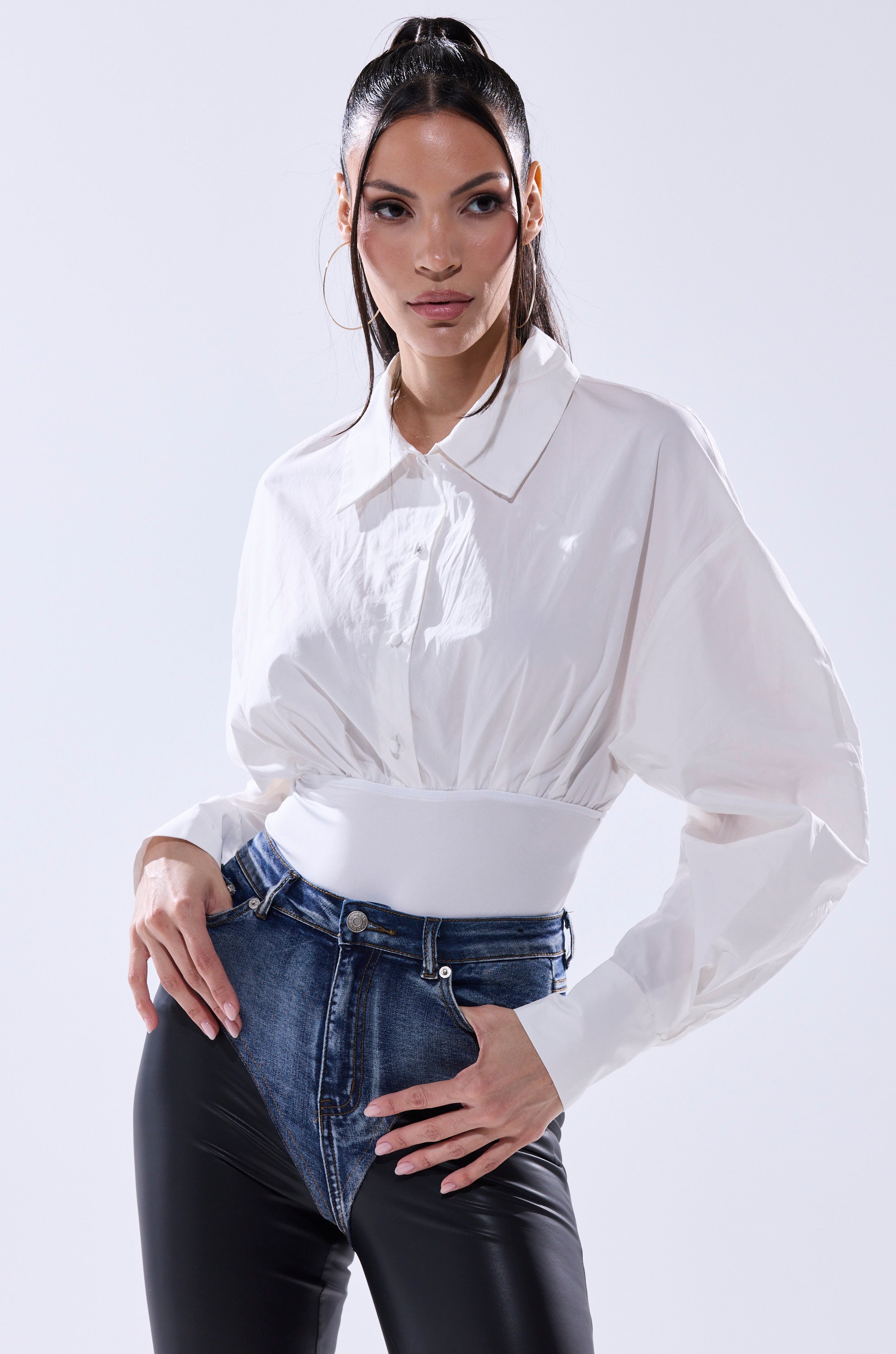 A woman poses confidently in the LET'S GO BABY BODYSUIT, a collared white long-sleeve crop top-pants set with a high waist and a faux leather belt, against a light background.