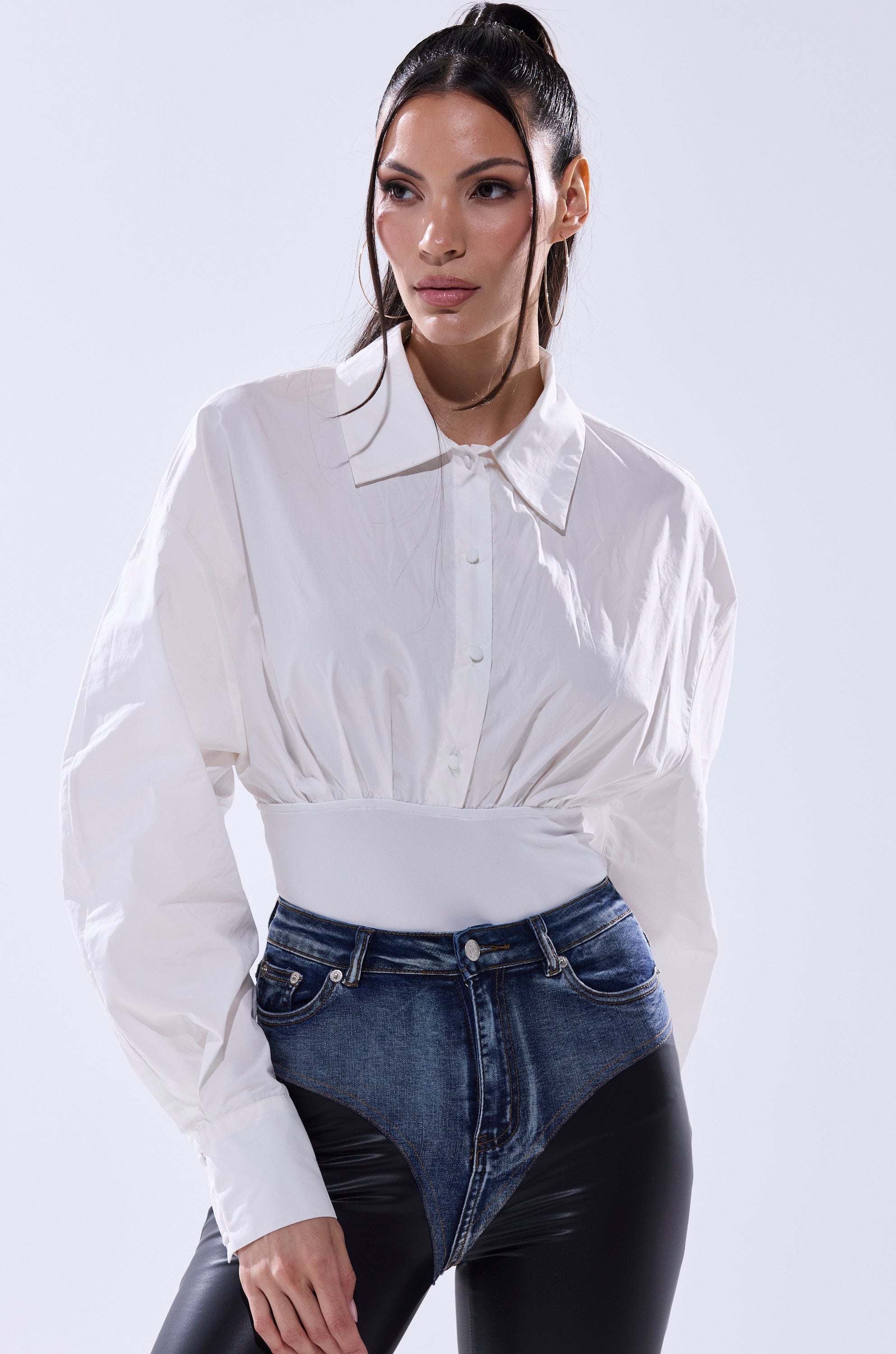A woman with long dark hair in a ponytail confidently poses in the LET'S GO BABY BODYSUIT, paired with a wide faux leather belt and high-waisted blue jeans layered over black pants against a plain background.