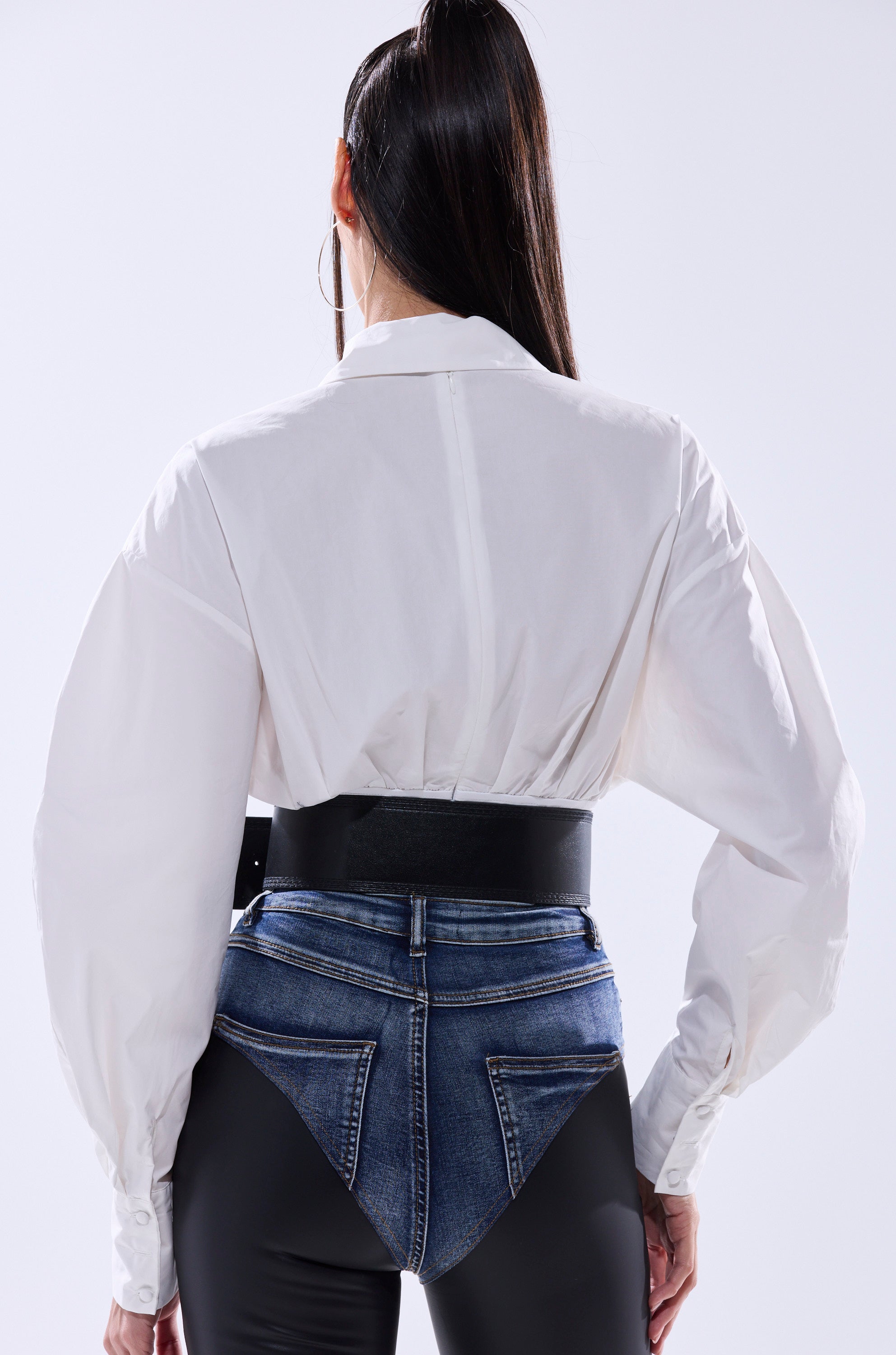 A person with long hair tied back stands against a plain background, wearing the LET'S GO BABY BODYSUIT in white with a faux leather belt and jeans wrapped below the hips over shiny black pants.