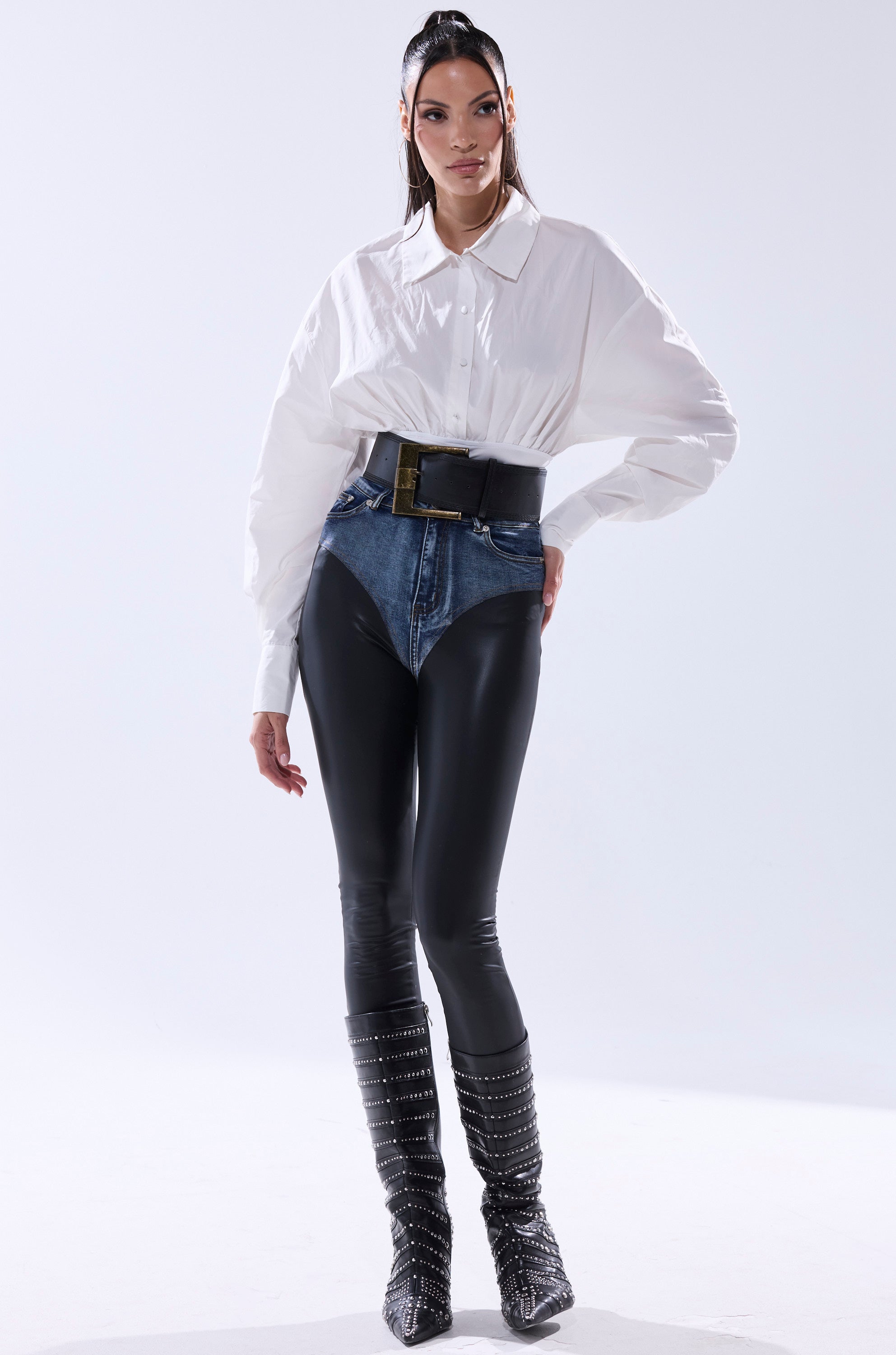 A woman poses in the LET'S GO BABY BODYSUIT, high-waisted denim, black leather-look pants, a faux leather belt, and knee-high studded black boots. Her long dark hair is styled in a high ponytail.