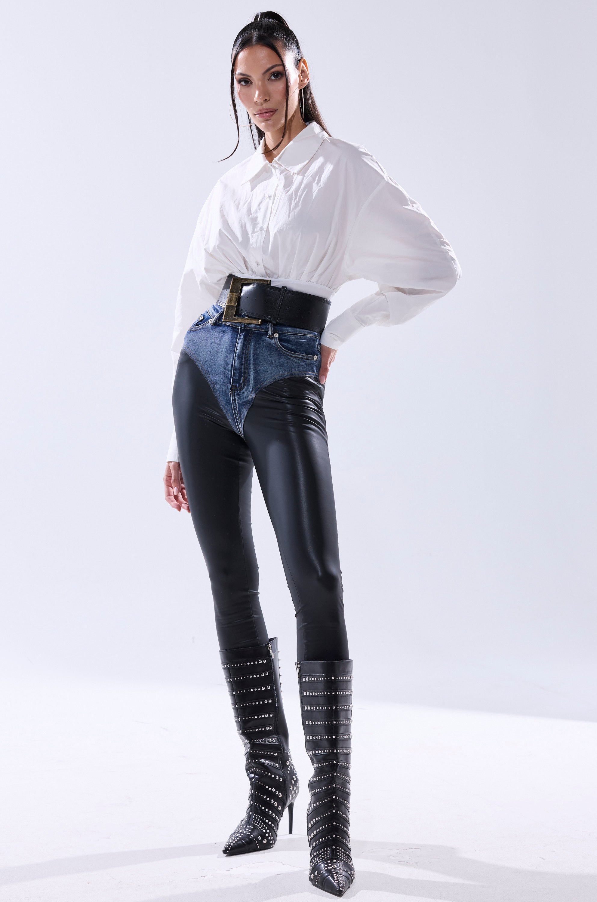 A woman poses confidently in the LET'S GO BABY BODYSUIT, styled with two-tone high-waisted pants, a faux leather belt, and knee-high black boots with silver studs. Her sleek hair stands out against a plain, well-lit background.