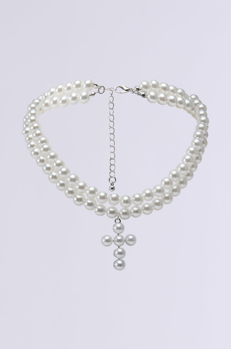 The PEARL DREAMING CROSS CHOKER is an exquisite three-strand necklace adorned with pearly beads and a hanging cross pendant. This layered accessory features a silver chain clasp for adjustable fitting, beautifully contrasted against a plain light gray background.