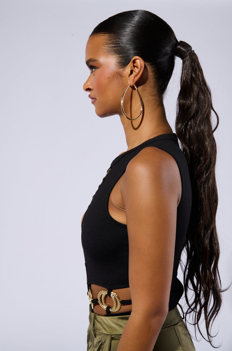 Profile view of a woman with long, sleek, dark hair tied in a high ponytail. She is wearing large hoop earrings, an olive green BRING IT ON CUT OUT BODYSUIT with a gold chain belt, and stretchy knit black pants. The background is plain light gray.