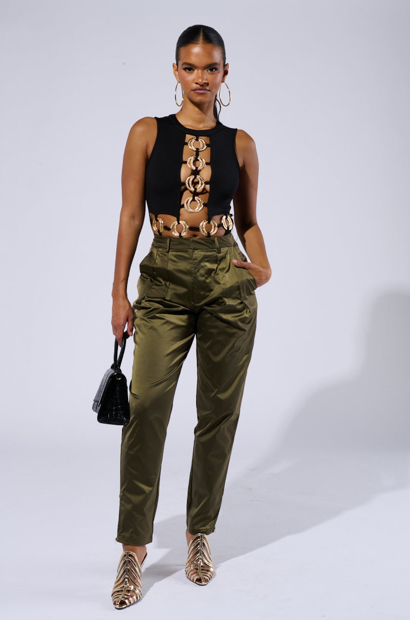 A woman stands confidently against a plain background wearing the BRING IT ON CUT OUT BODYSUIT, featuring metallic ring details and sexy paneled cut-outs. She pairs it with olive green high-waisted pants and animal-print pointed flats. She holds a small black handbag, accessorized with hoop earrings, and has her hair slicked back.