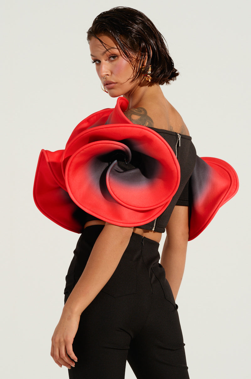 A person with short hair showcases a striking ensemble, wearing the PRETTY GIRL ROSE TOP adorned with a large red and black rose and sculptural sleeve detail over one shoulder. They pair it with black high-waisted pants against a plain background.