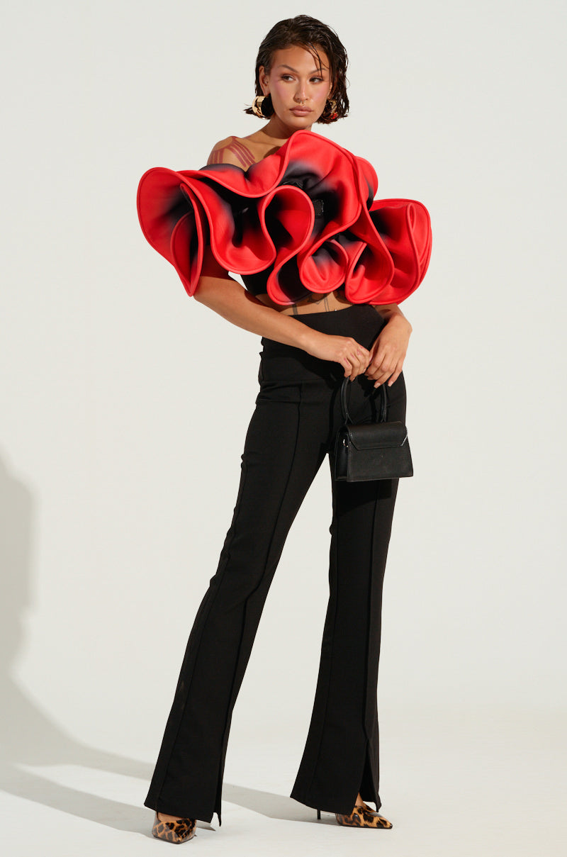 A person stands confidently wearing the PRETTY GIRL ROSE TOP featuring ruffle detailing, paired with black pants. They hold a small black handbag and wear leopard-print shoes, while the minimalist, light-colored backdrop highlights their striking ensemble.