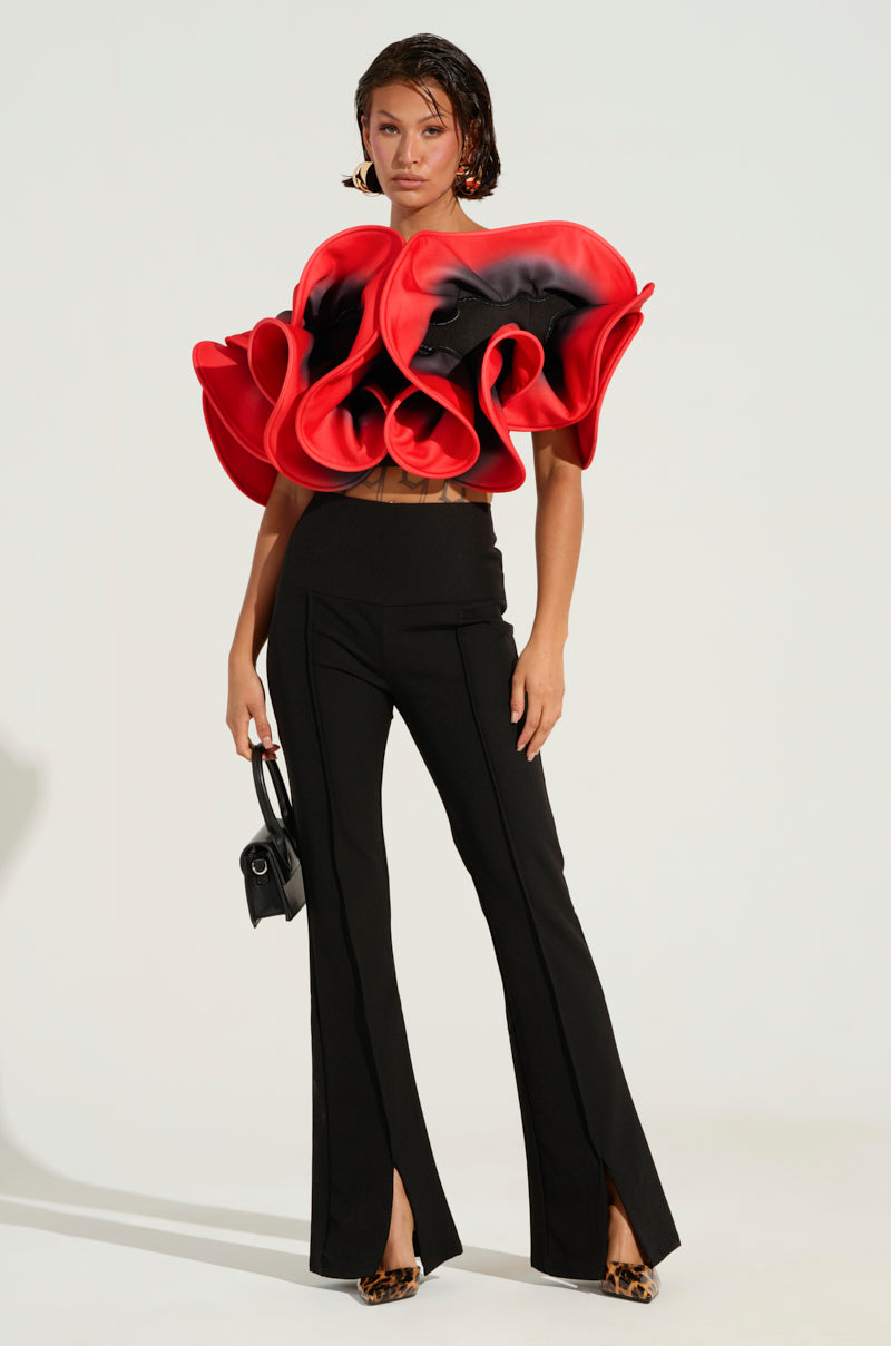 A woman poses in a bold fashion ensemble featuring the PRETTY GIRL ROSE TOP, characterized by its striking ruffle detailing, paired with sleek black pants. She accessorizes with a small handbag and styles her hair in a trendy wet look. The outfit is finished off with leopard-print shoes.