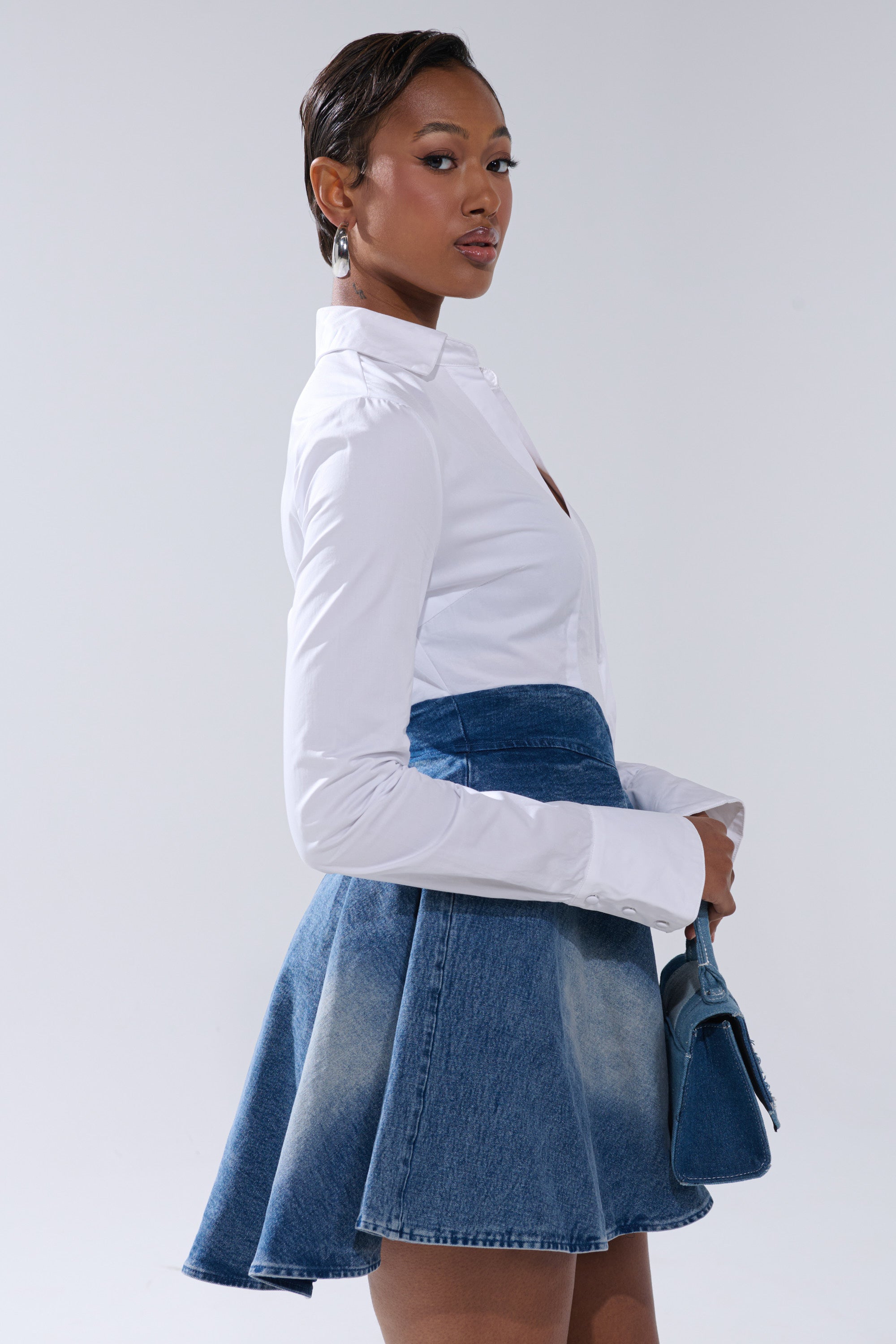 A woman stands sideways, confidently posing in the DONT NEED NOBODY DENIM MINI DRESS and holding a small blue purse against a plain light background.