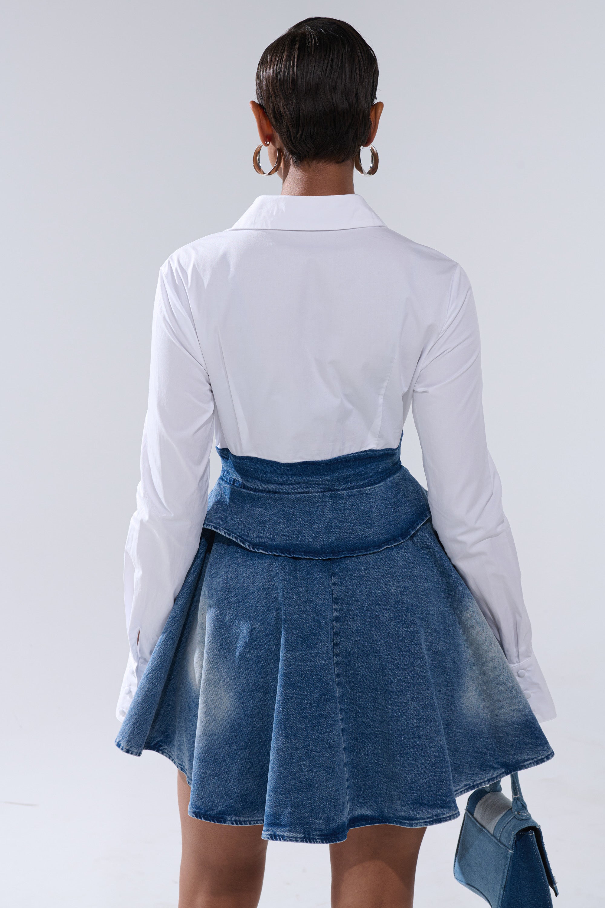 Wearing the DONT NEED NOBODY DENIM MINI DRESS, a person with short, slicked-back hair and large hoop earrings stands facing away, holding a small denim handbag and pairing the dress with a white button-down shirt underneath.