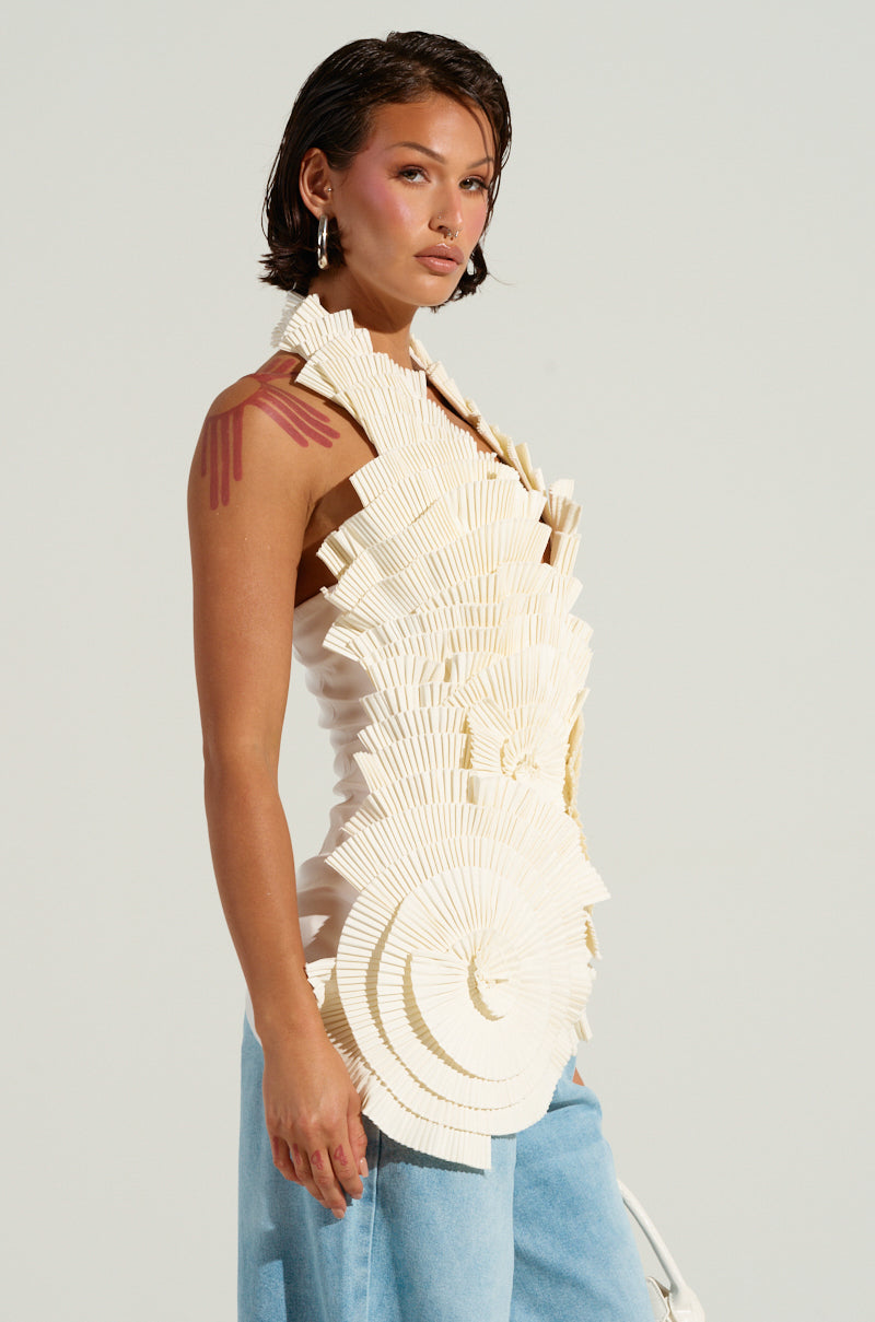 A person with short brown hair wears the distinctive COVER ME IN LOVE white faux leather top, characterized by halter neck straps and an asymmetrical hem that resembles layered paper fans. They pair it with blue pants and stand confidently against a plain background, subtly showcasing a tattoo on their left arm.