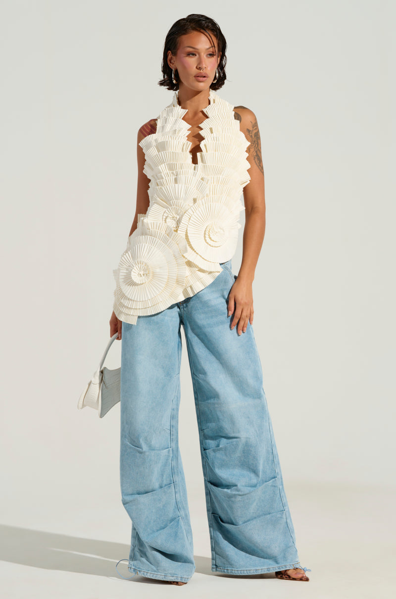A woman wears the COVER ME IN LOVE TOP, a sleeveless white top adorned with intricate circular ruffle patterns and an asymmetrical hem, paired with wide-leg blue jeans. She carries a small white bag while standing against a plain background, gazing to the side.