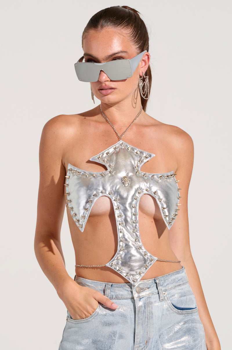 A person wears oversized sunglasses and the WATCHING OVER ME CROSS TOP, a metallic silver halter neck adorned with skull embellishments and featuring an open chest design. They pair it with light blue jeans against a plain, neutral background for an edgy fashion statement.