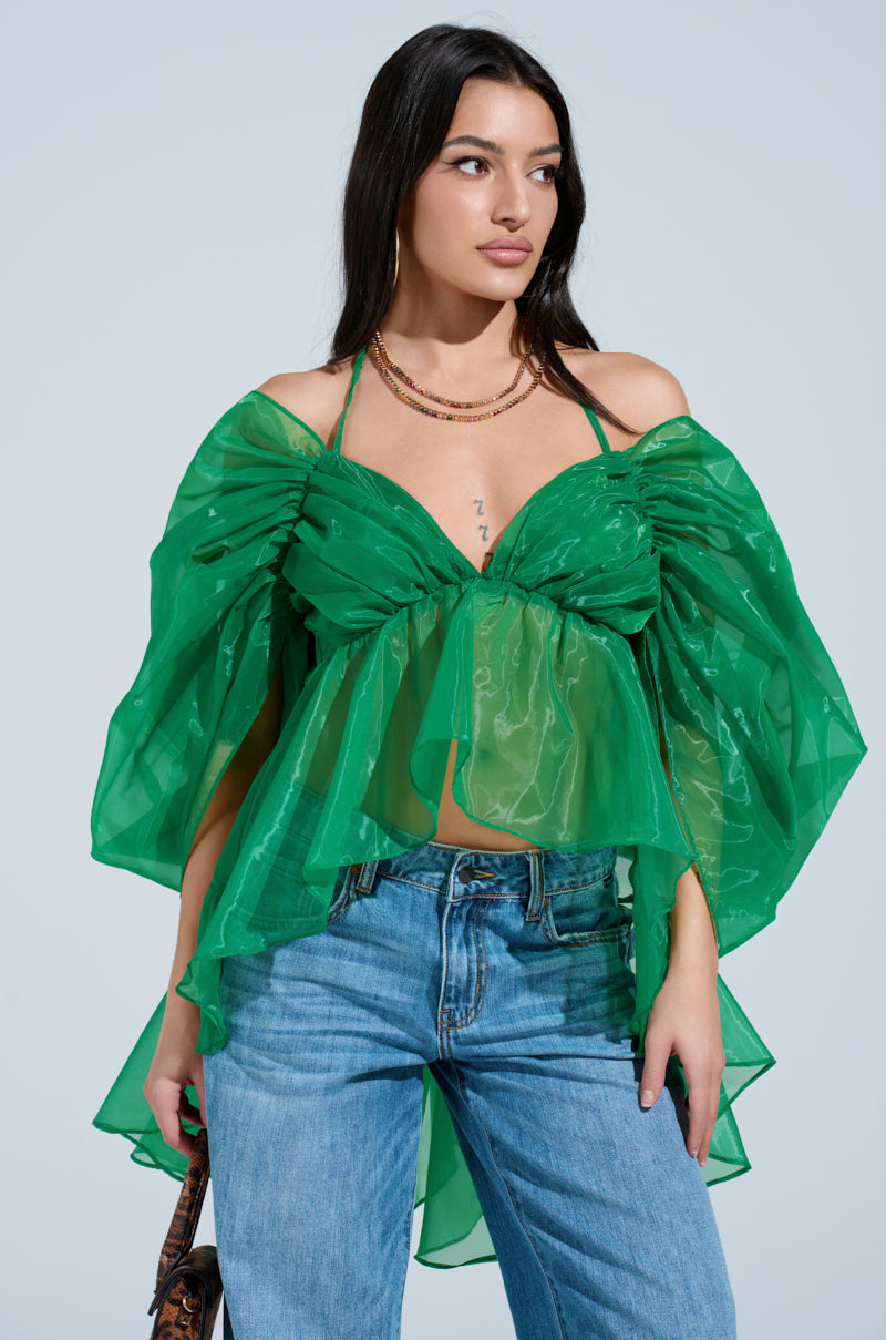 A woman wears the AN ELEGANT EVENING ORGANZA BLOUSE in sheer Kelly Green with dramatic ruffled sleeves and blue jeans. She has long dark hair, layered necklaces, and poses against a light blue background.