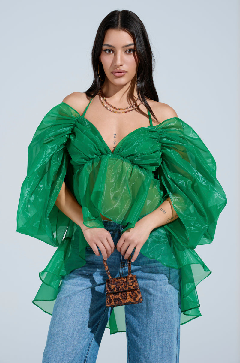 A woman with long dark hair wears AN ELEGANT EVENING ORGANZA BLOUSE in kelly green with sheer, voluminous off-shoulder sleeves, paired with blue jeans, gold hoop earrings, layered necklaces, and a small brown snakeskin handbag.